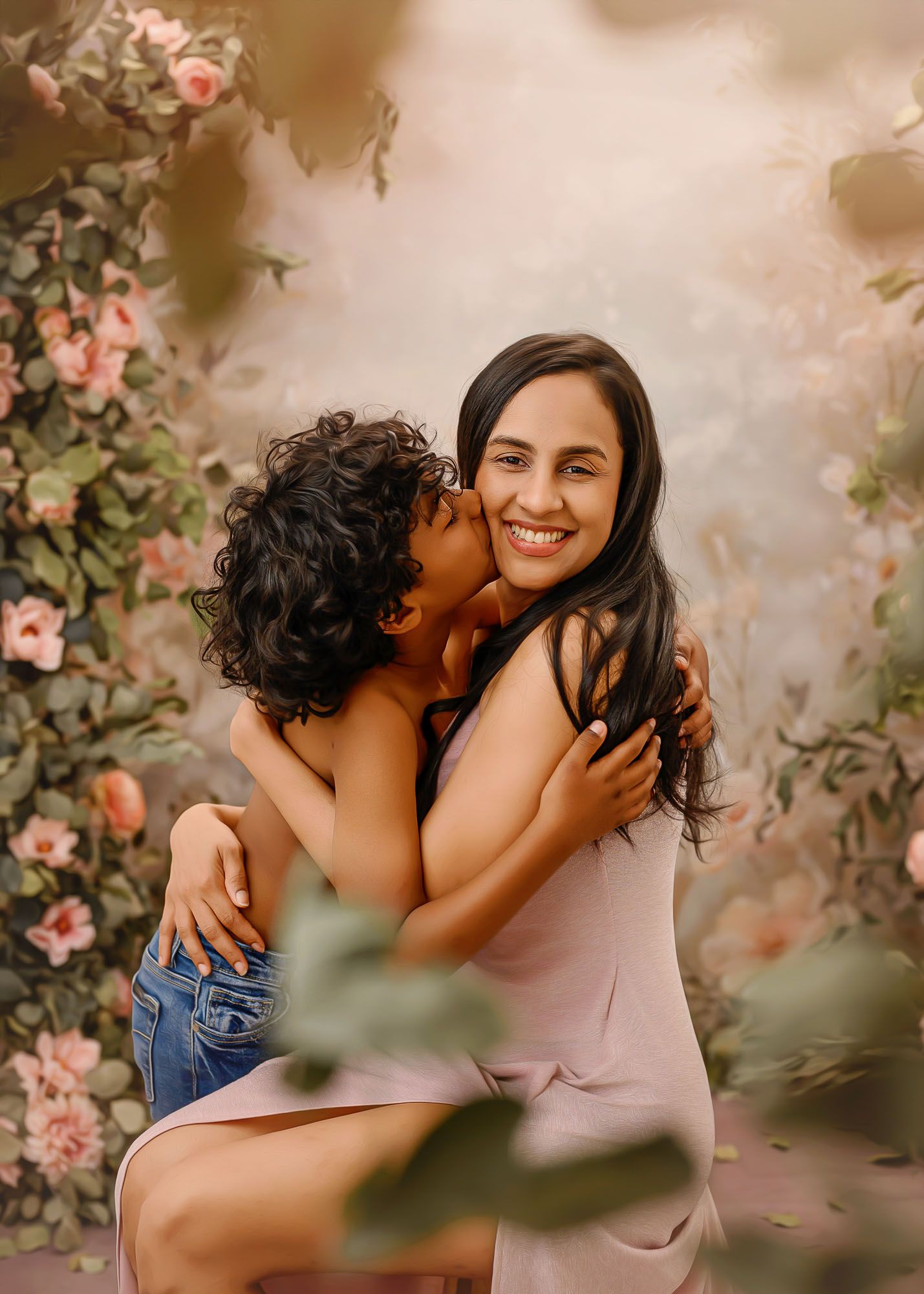 A mother smiles joyfully as her curly-haired child hugs and kisses her cheek amid a floral backdrop.