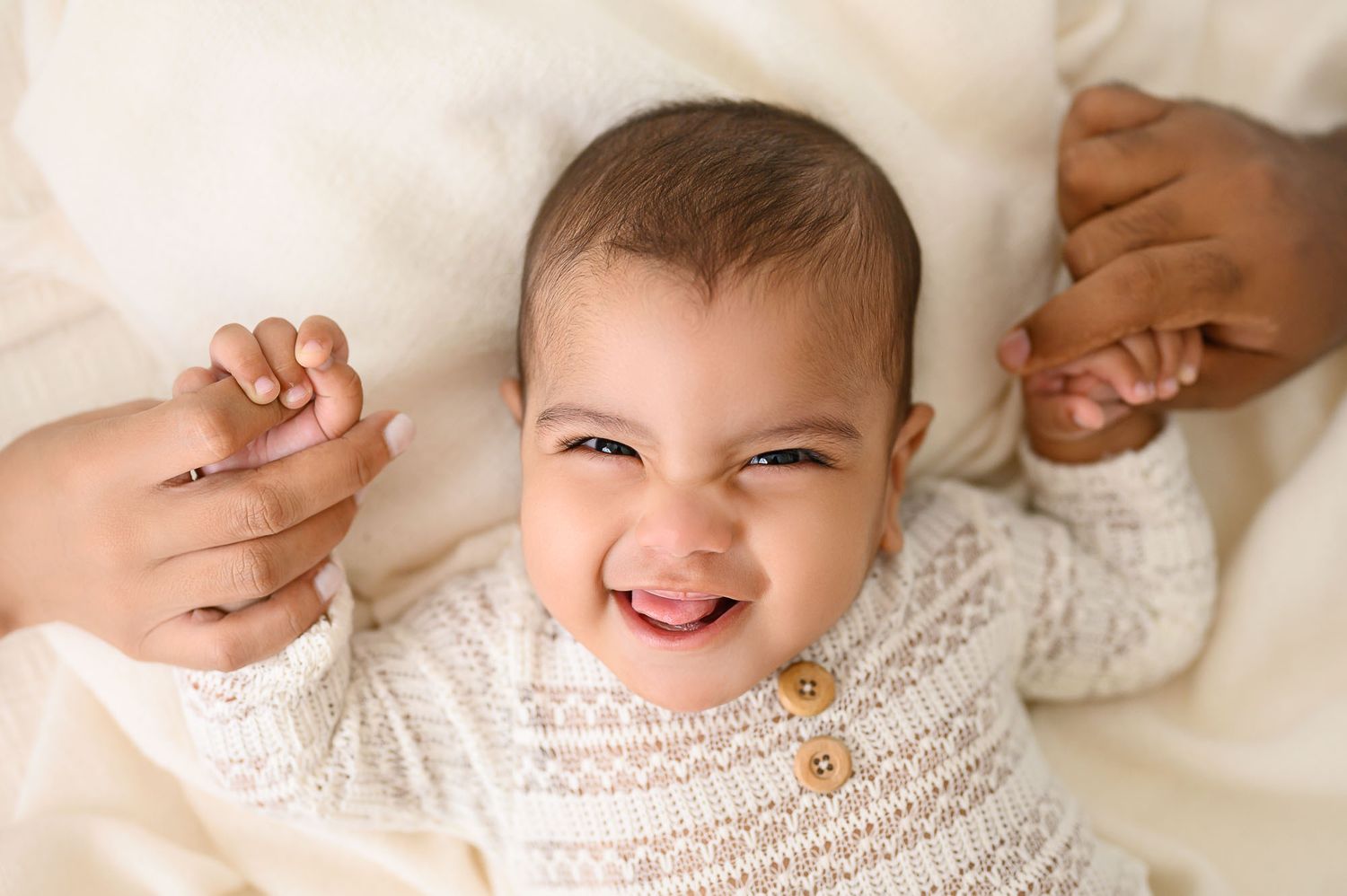 Smiling baby in cream knit outfit held by parents hands on soft white blanket.