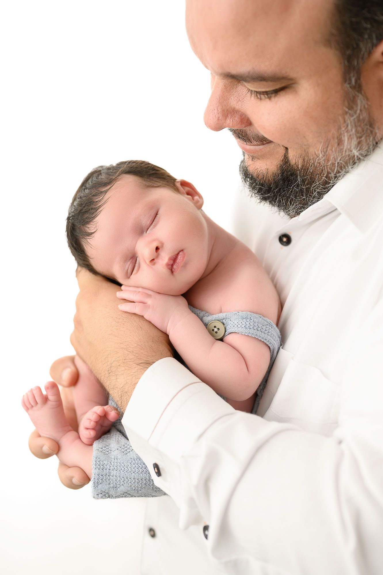 Bearded father in white shirt smiling while holding sleeping newborn baby dressed in blue.