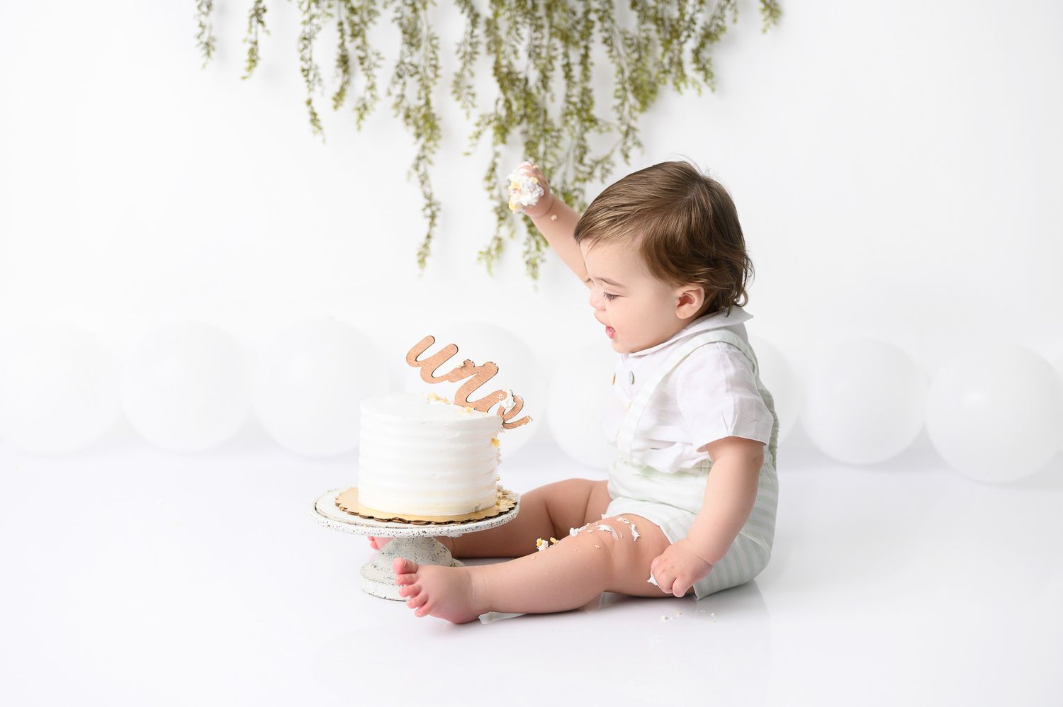 Baby in white outfit reaching for cake during cake smash photo session.