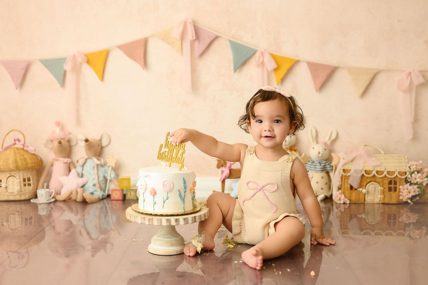 Baby girl in yellow romper touches first birthday cake surrounded by bunny toys and pastel pennant banners.