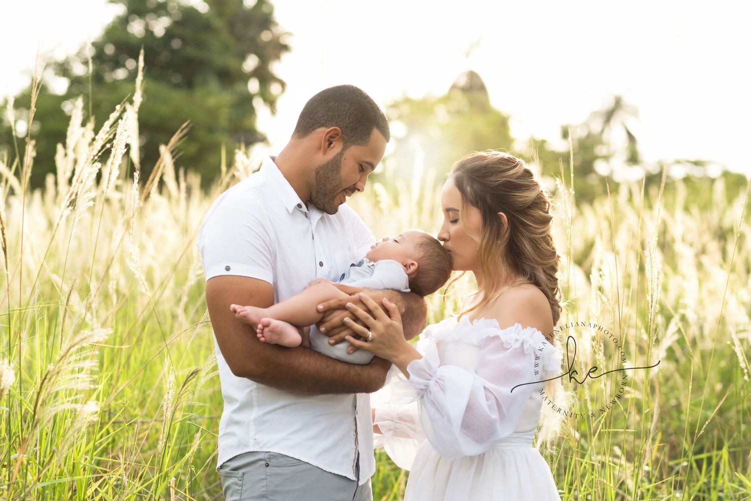 A couple stands in a sunlit field holding their newborn baby, surrounded by tall grass in a golden outdoor setting.