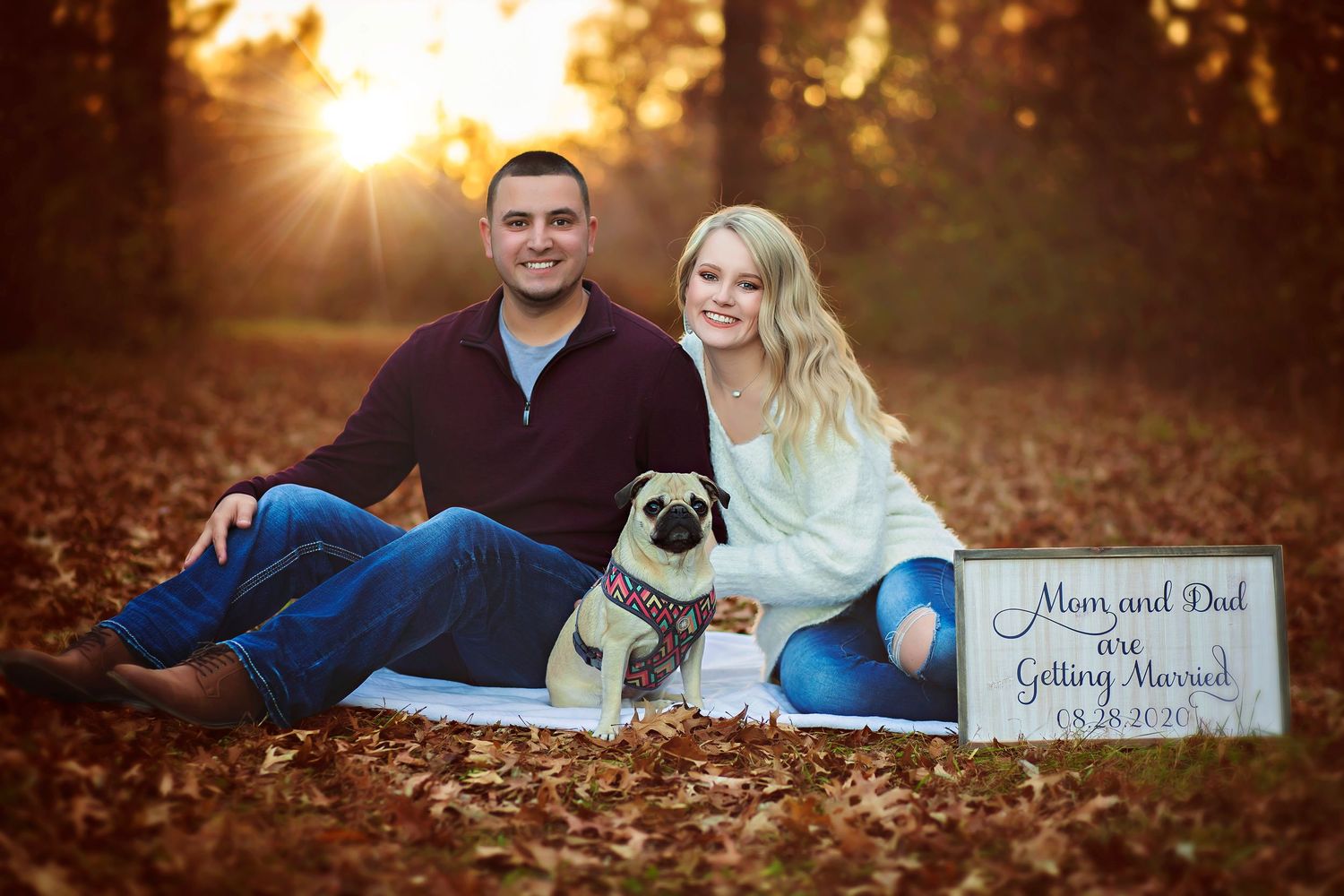 Couple sitting in autumn leaves with pug dog and save the date sign announcing wedding date 09.28.2019.
