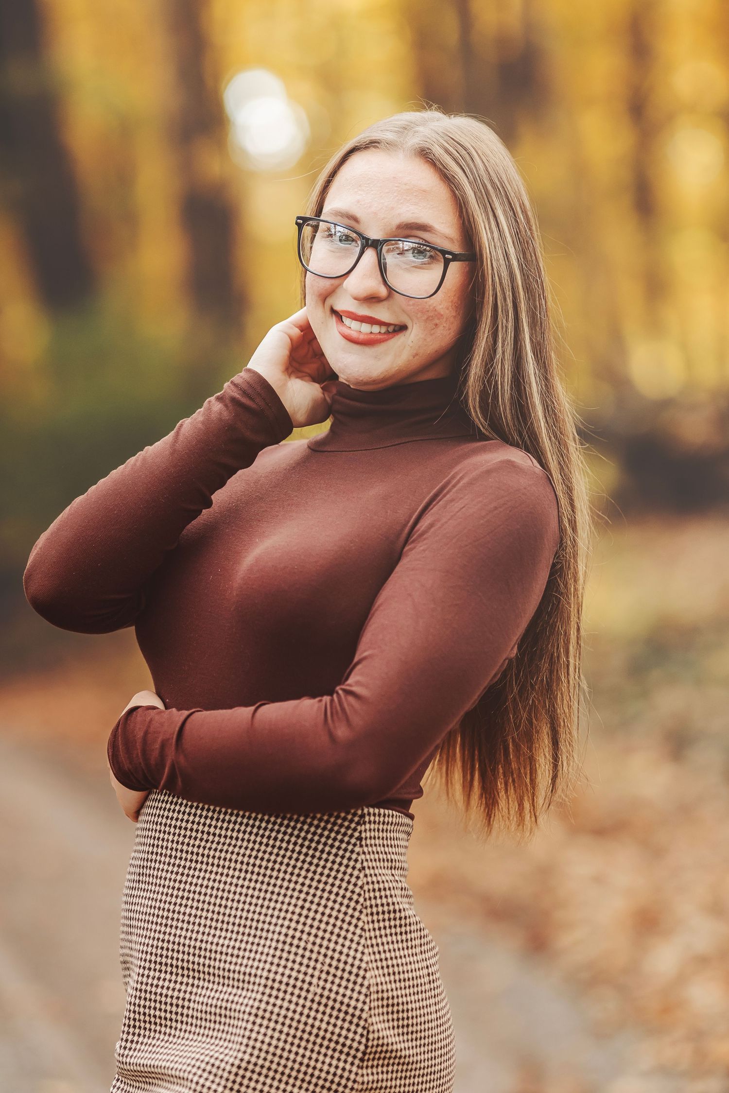Young woman with glasses and long hair smiles in autumn forest, wearing brown turtleneck and plaid skirt.