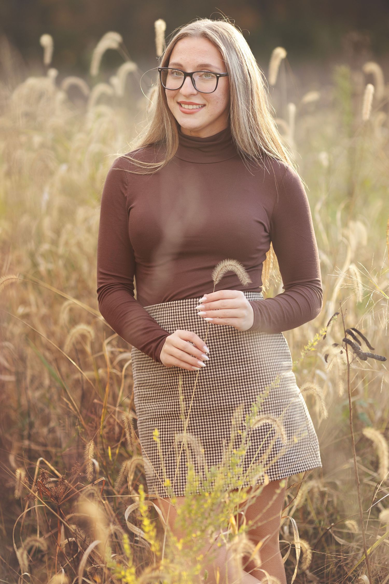 Smiling blonde woman in glasses and brown turtleneck with plaid skirt standing in a golden field.