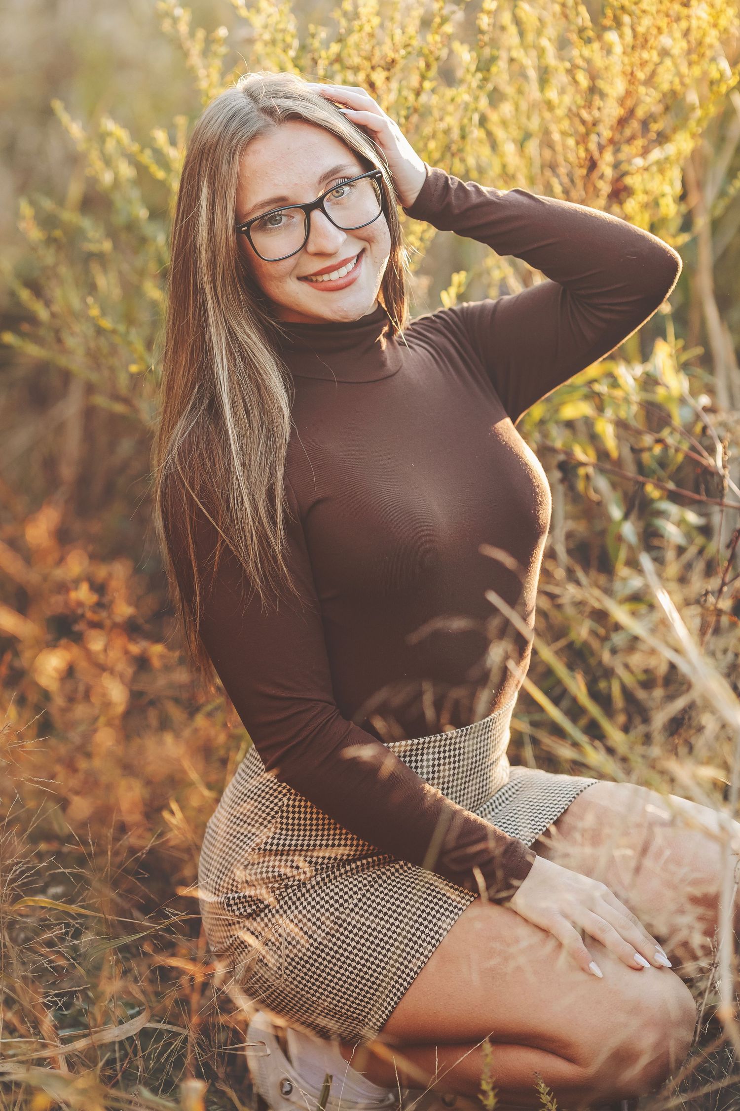 Smiling young woman with glasses and long blonde hair sitting outdoors in autumn field wearing brown turtleneck and plaid skirt.