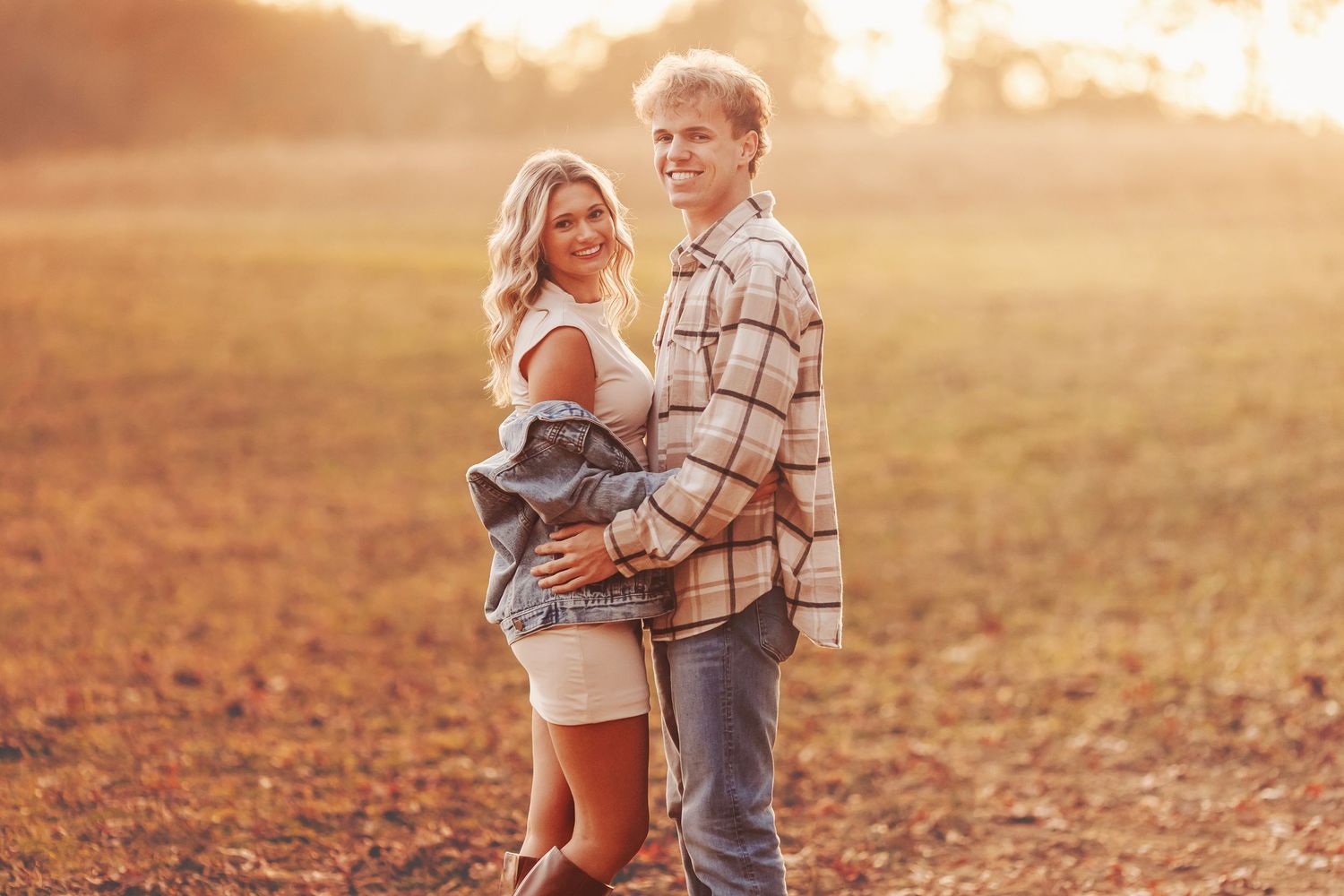 Couple posing together in a sunlit field during golden hour, wearing casual plaid and denim outfits.