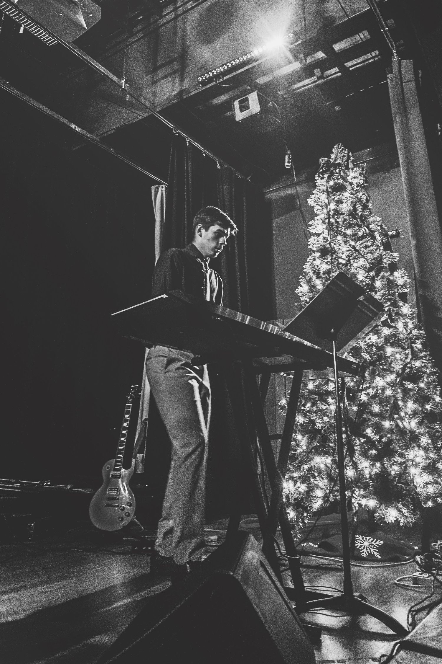 Musician playing keyboard on stage beside a lit Christmas tree in black and white photo.