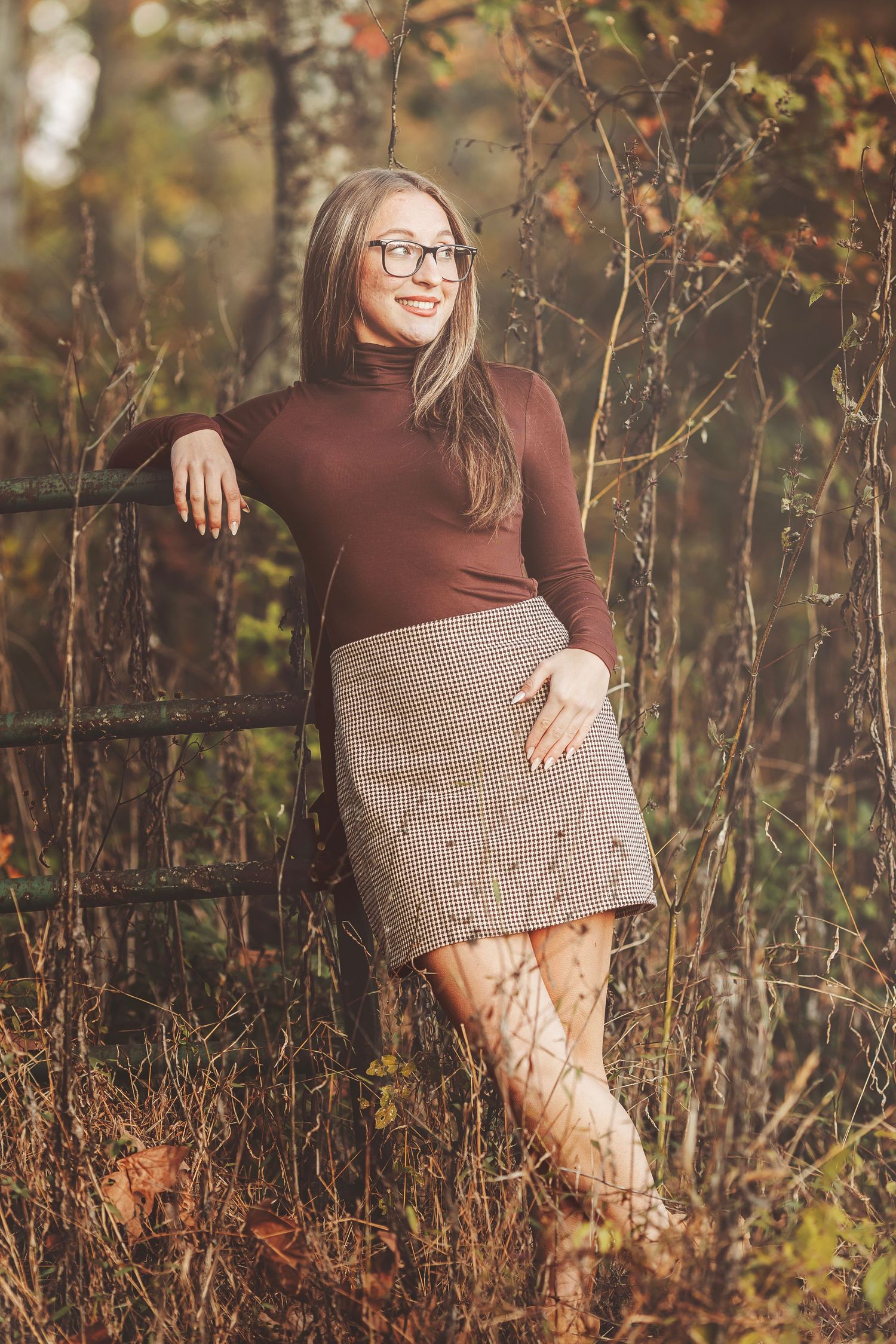 Woman in brown turtleneck and plaid skirt with glasses poses outdoors in autumn forest setting.