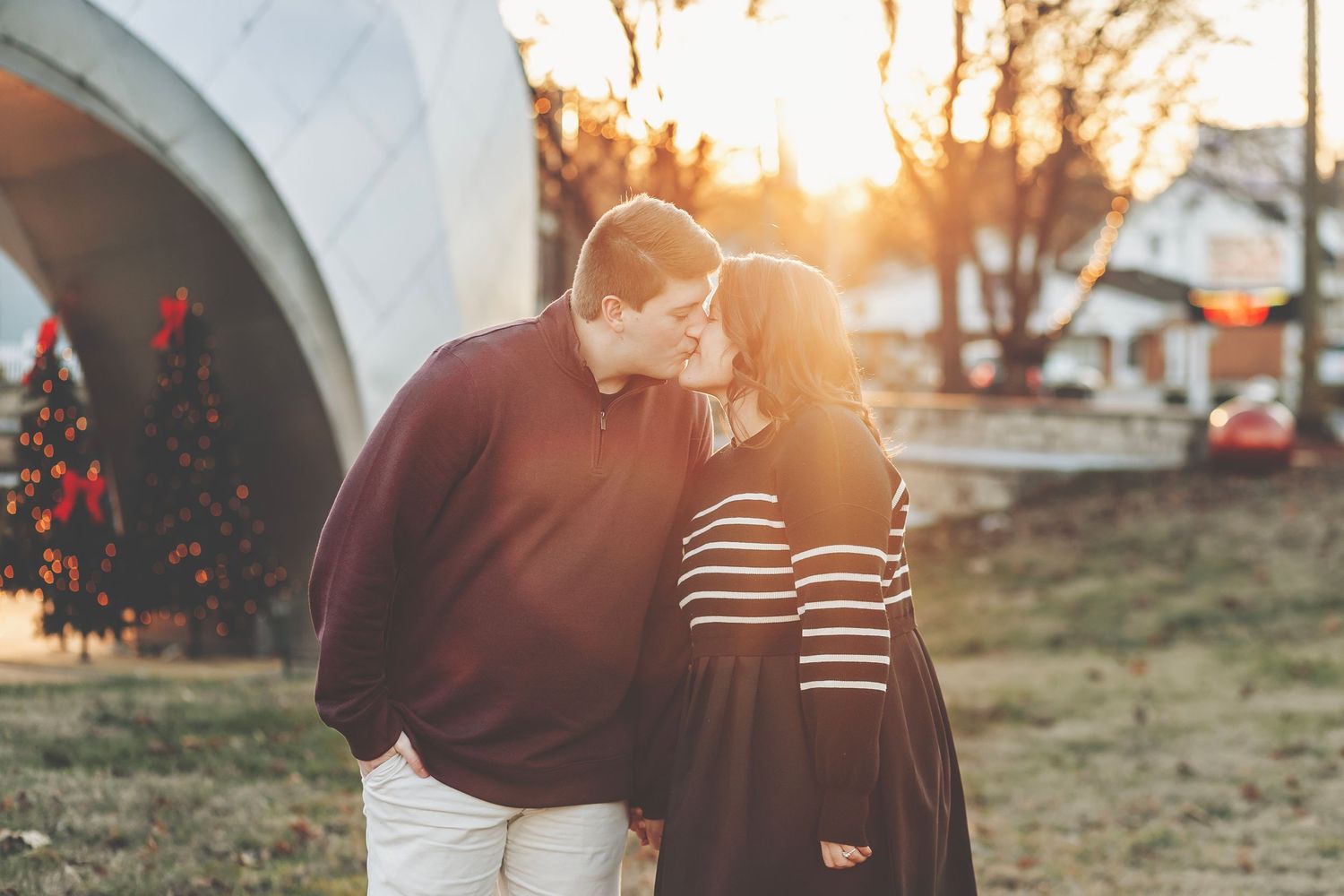 Couple sharing a tender kiss at sunset near a Christmas tree outdoors in a warm romantic setting.