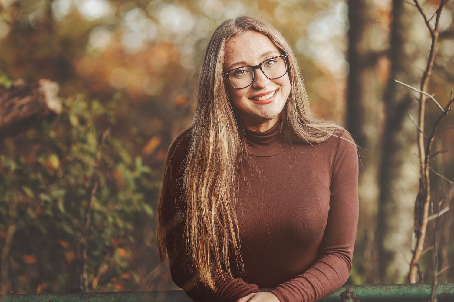 Young woman with long blonde hair and glasses smiling outdoors in autumn setting wearing brown turtleneck.