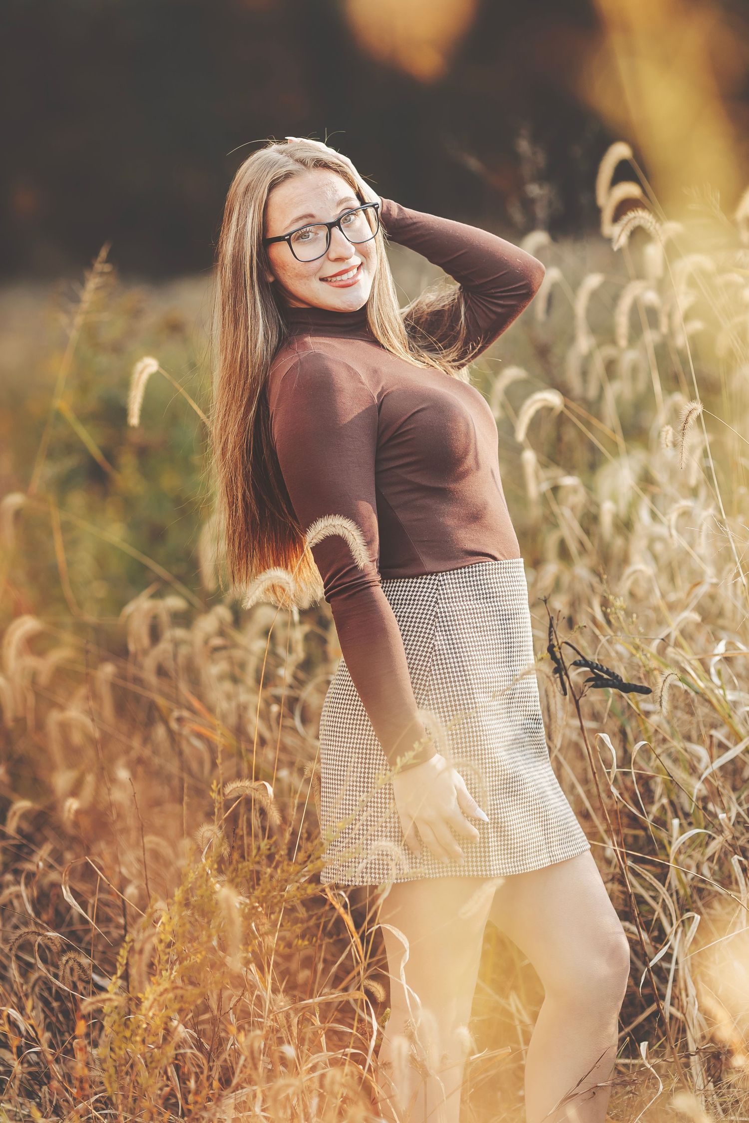 Young woman in brown turtleneck and plaid skirt posing in golden autumn field with tall grass.