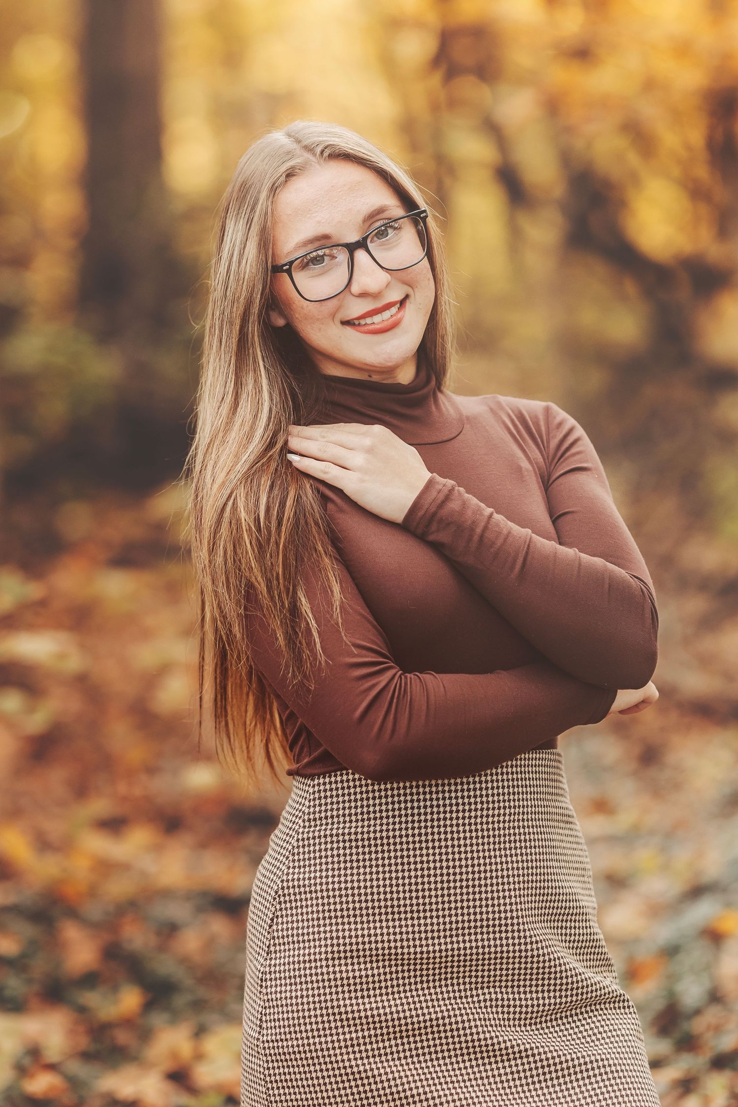 Young woman with long hair and glasses smiling in autumn forest, wearing brown turtleneck and plaid skirt.