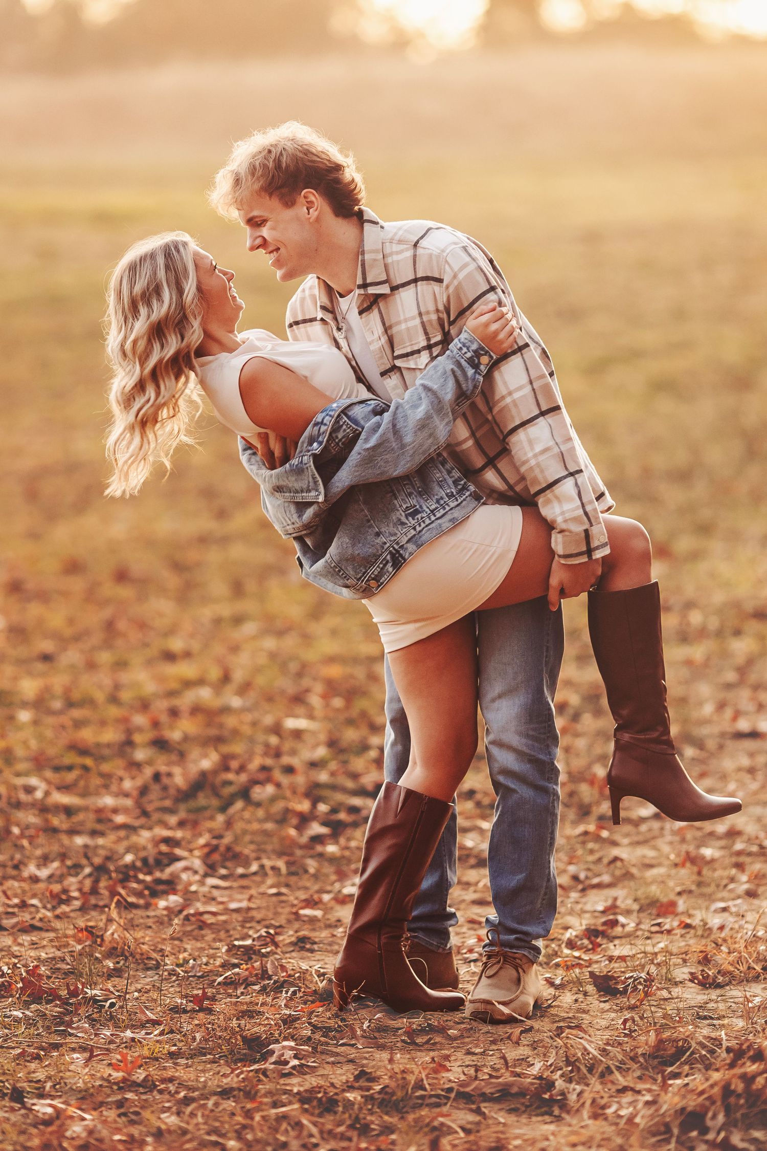 Couple dancing romantically in an autumn field at golden hour, woman in boots lifted by man in plaid shirt.