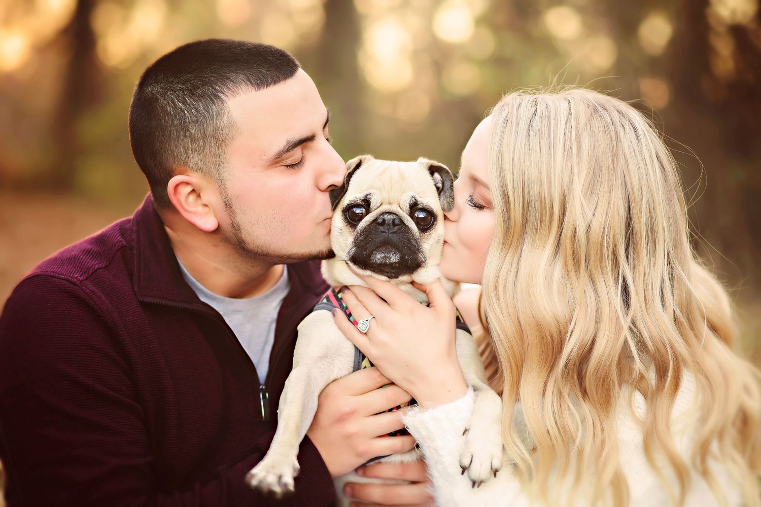Couple kissing a cute pug dog outdoors in a warm autumn setting with golden bokeh background.