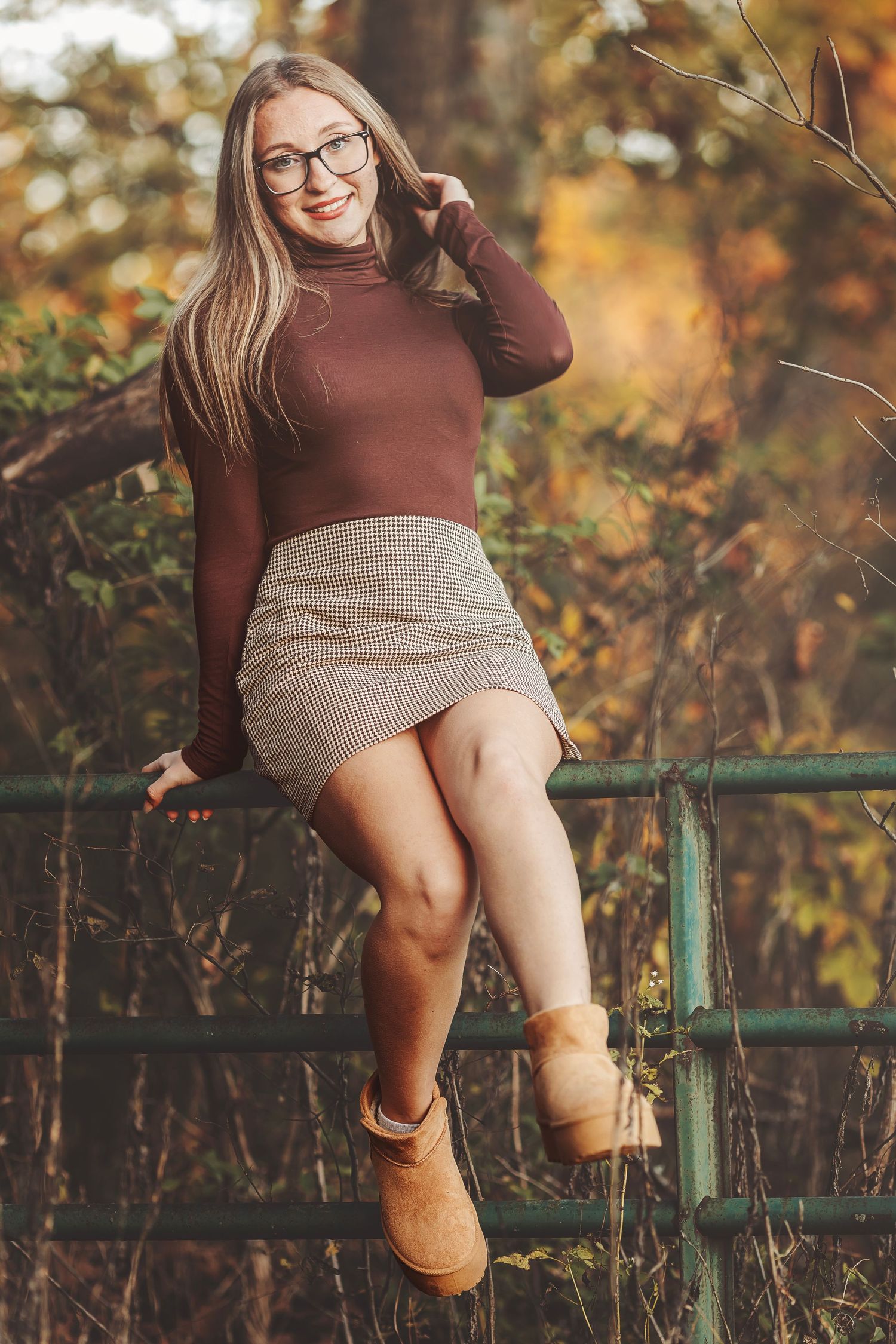 Smiling blonde woman in glasses sitting on rustic fence in colorful autumn forest setting.