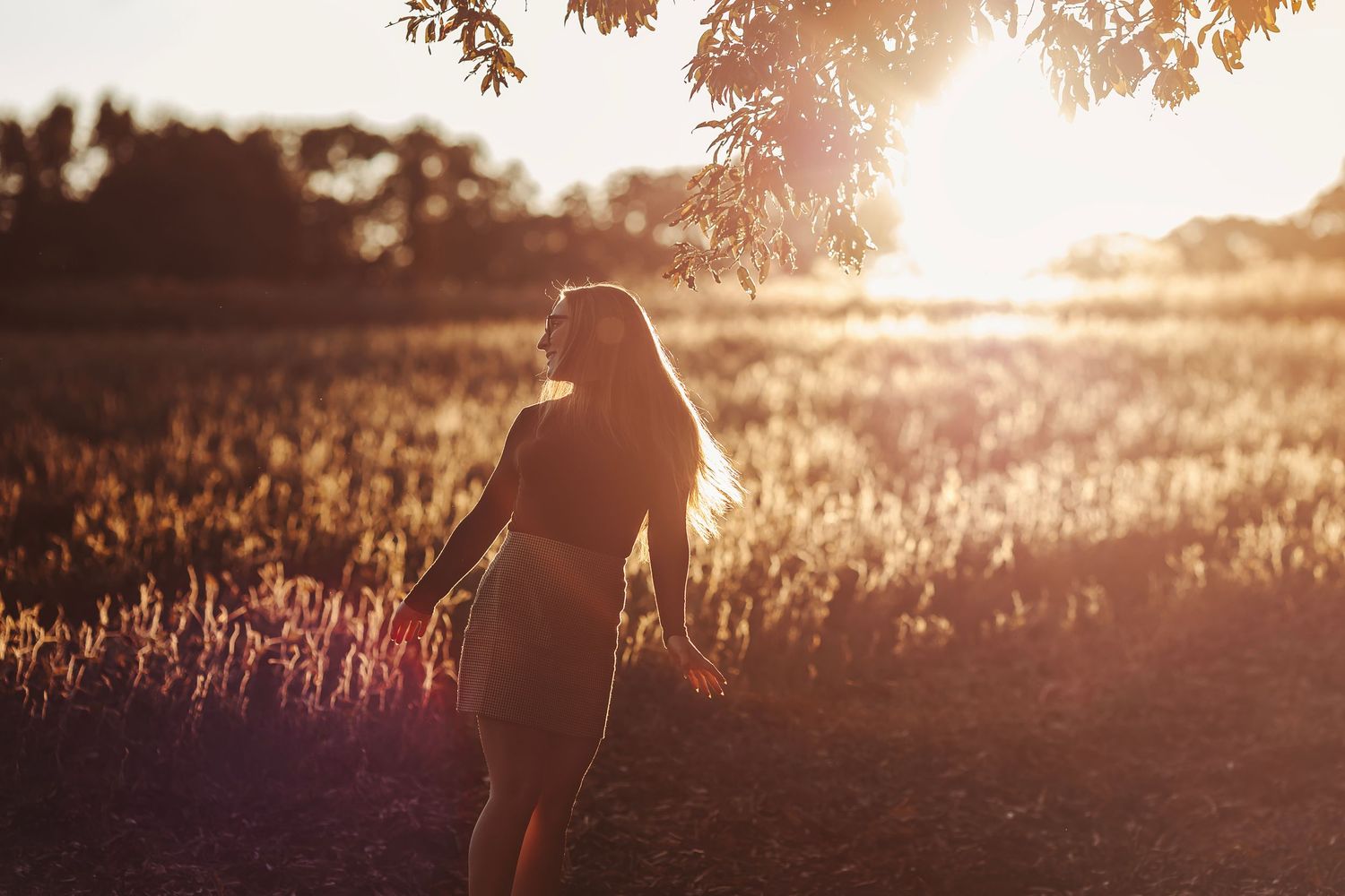 Woman standing in golden sunlit field at sunset with long hair blowing, surrounded by wildflowers and trees.