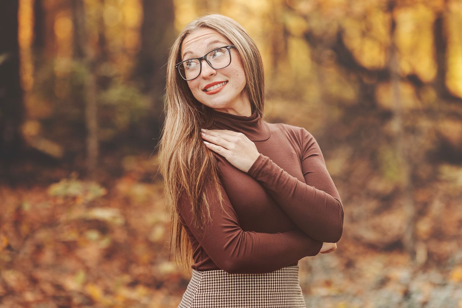 Young woman with glasses and long blonde hair smiling in autumn forest wearing brown turtleneck and plaid skirt.