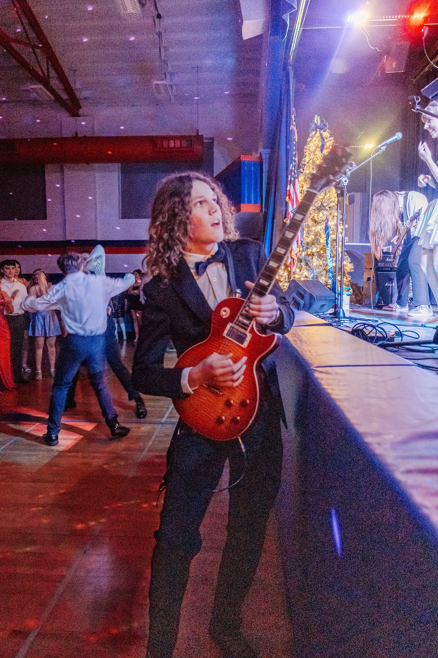 Guitarist in black tuxedo playing red Les Paul electric guitar at festive holiday party event with dancers in background.