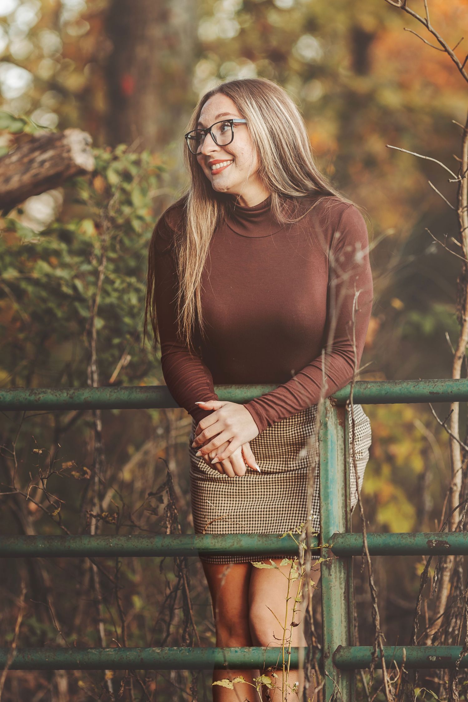 Smiling blonde woman in glasses, brown turtleneck, and plaid skirt leaning on a green fence in autumn.