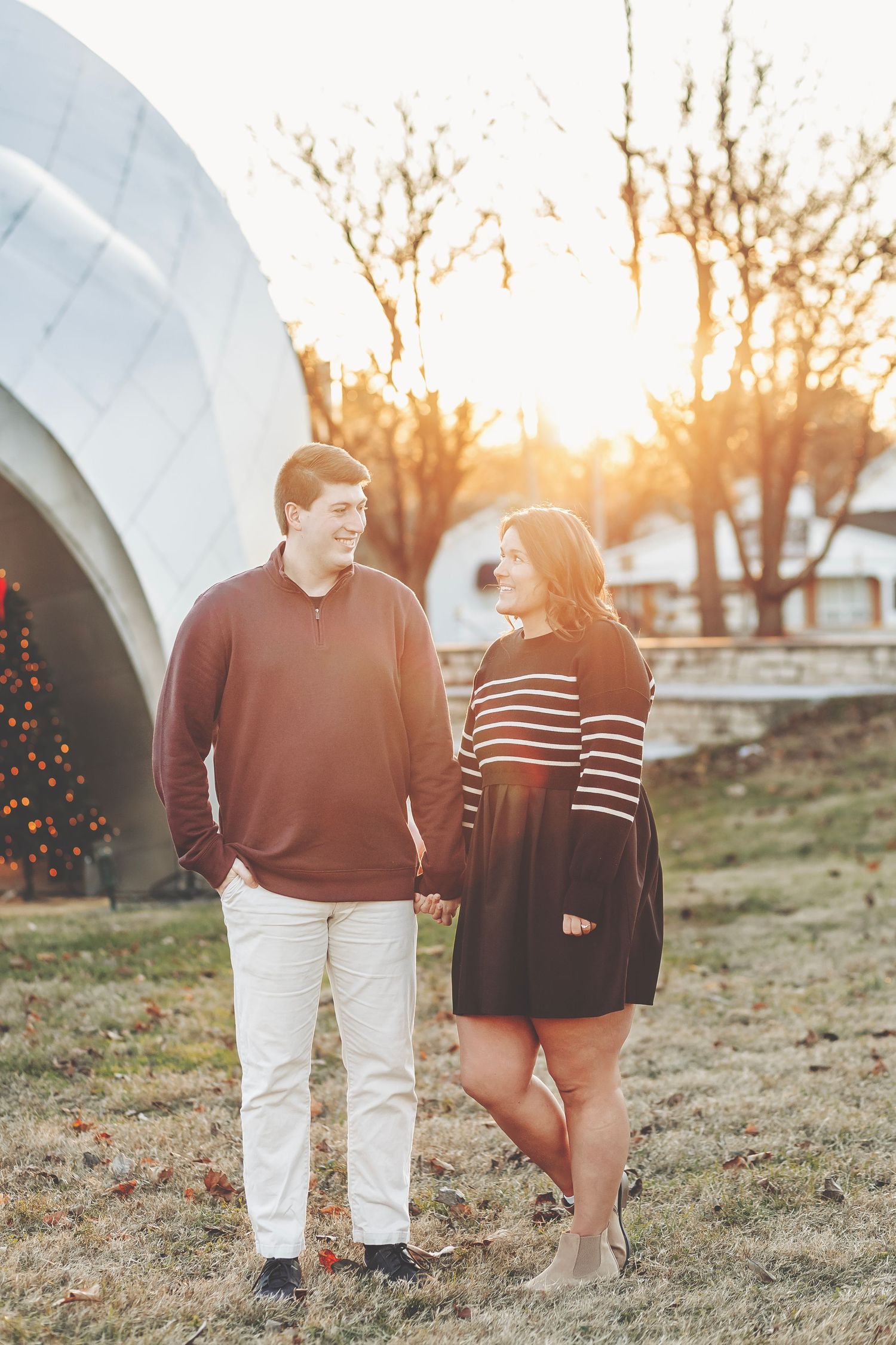 Couple holding hands at golden hour sunset outdoors near a dome structure and Christmas tree.