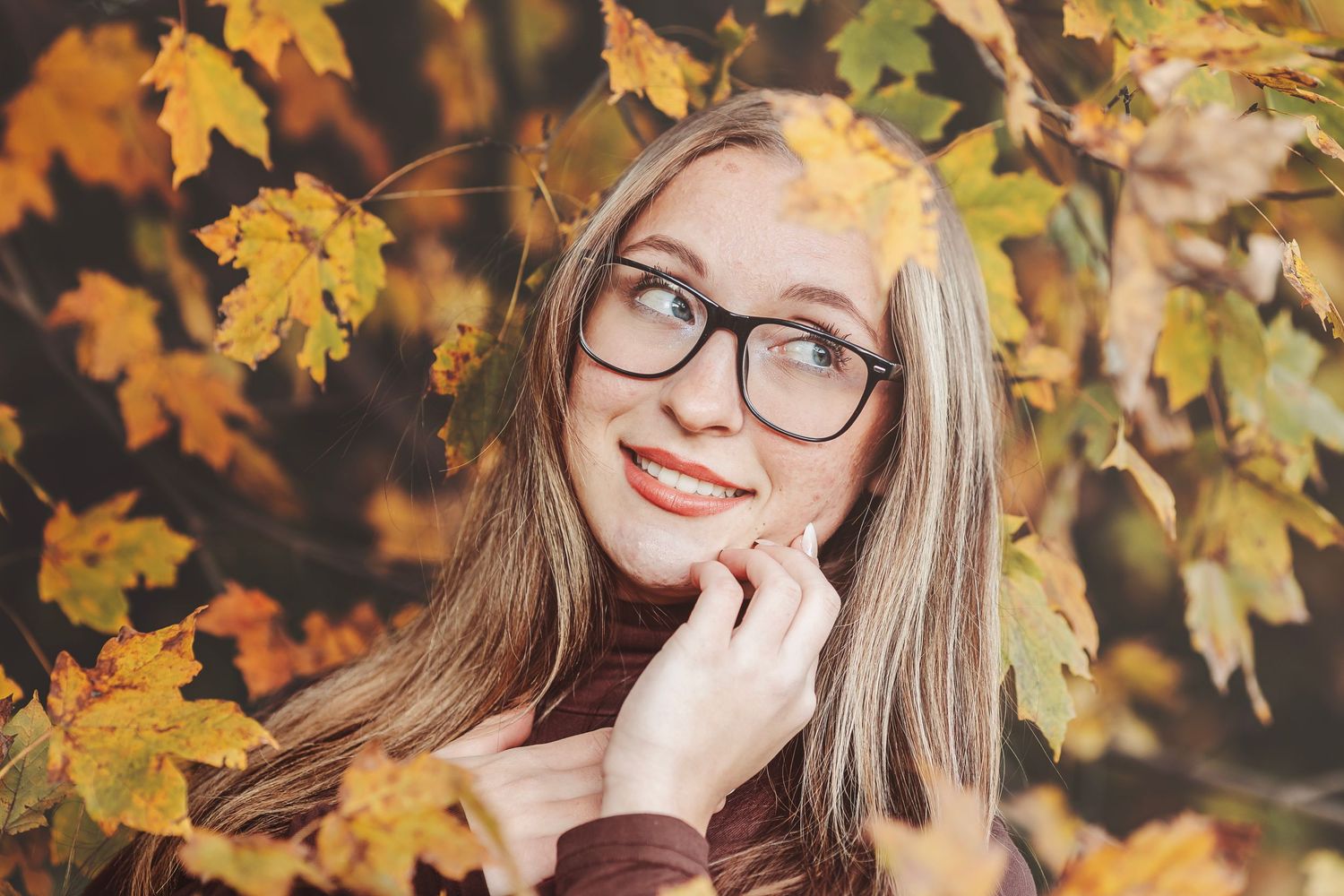 Young woman with glasses smiling among vibrant autumn leaves in a warm fall outdoor portrait.