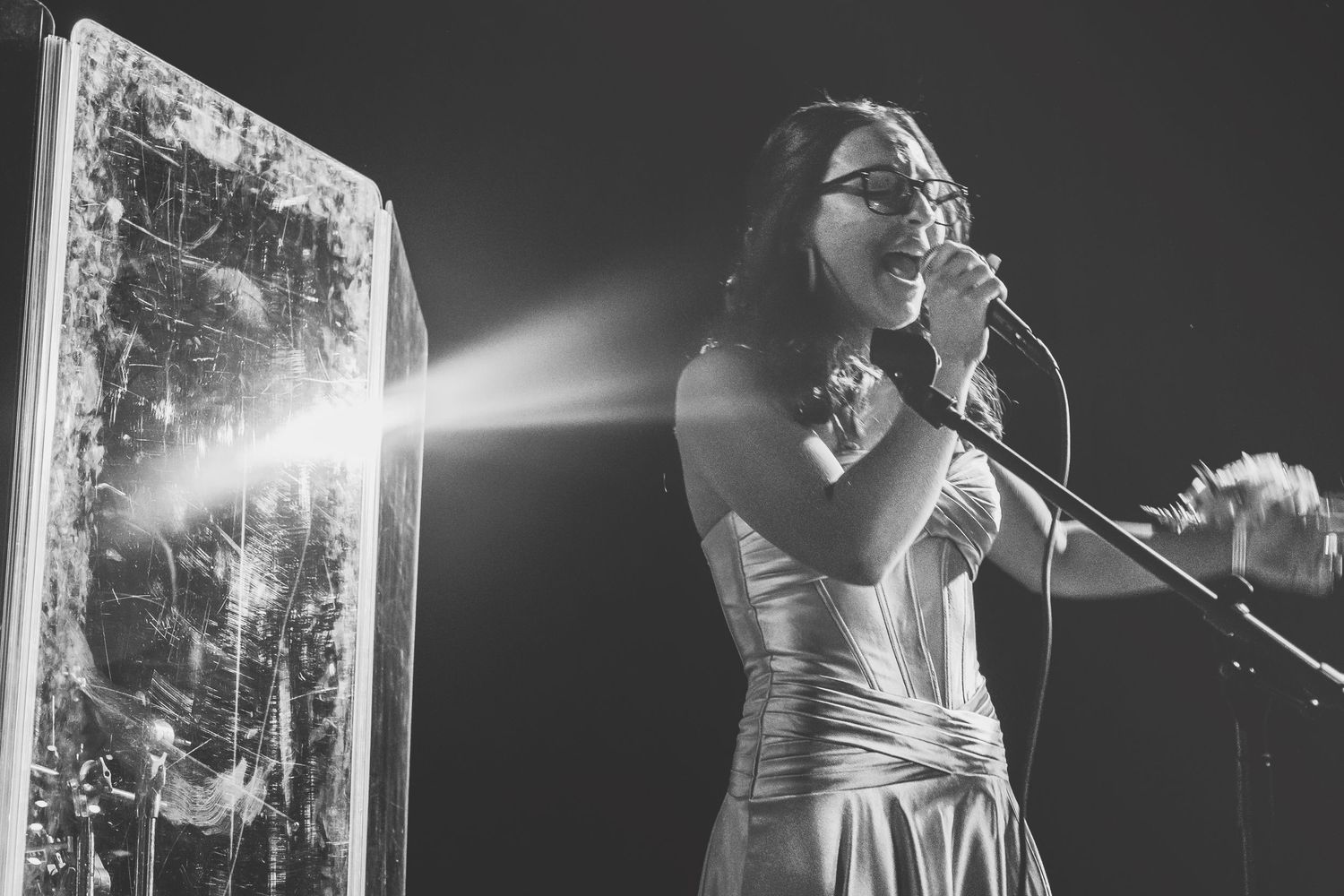 Black and white photo of a woman singing into a microphone on stage with dramatic light beam effect.