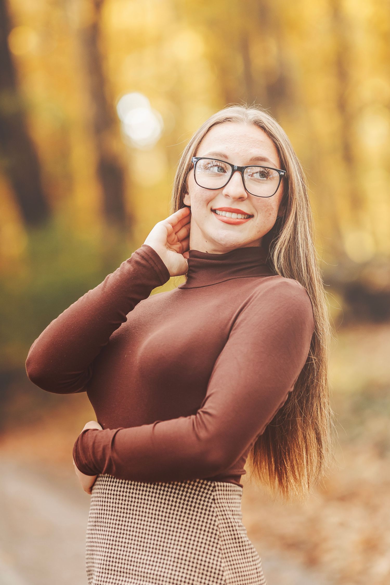 Young woman with glasses and brown turtleneck smiling in autumn forest with golden foliage background.