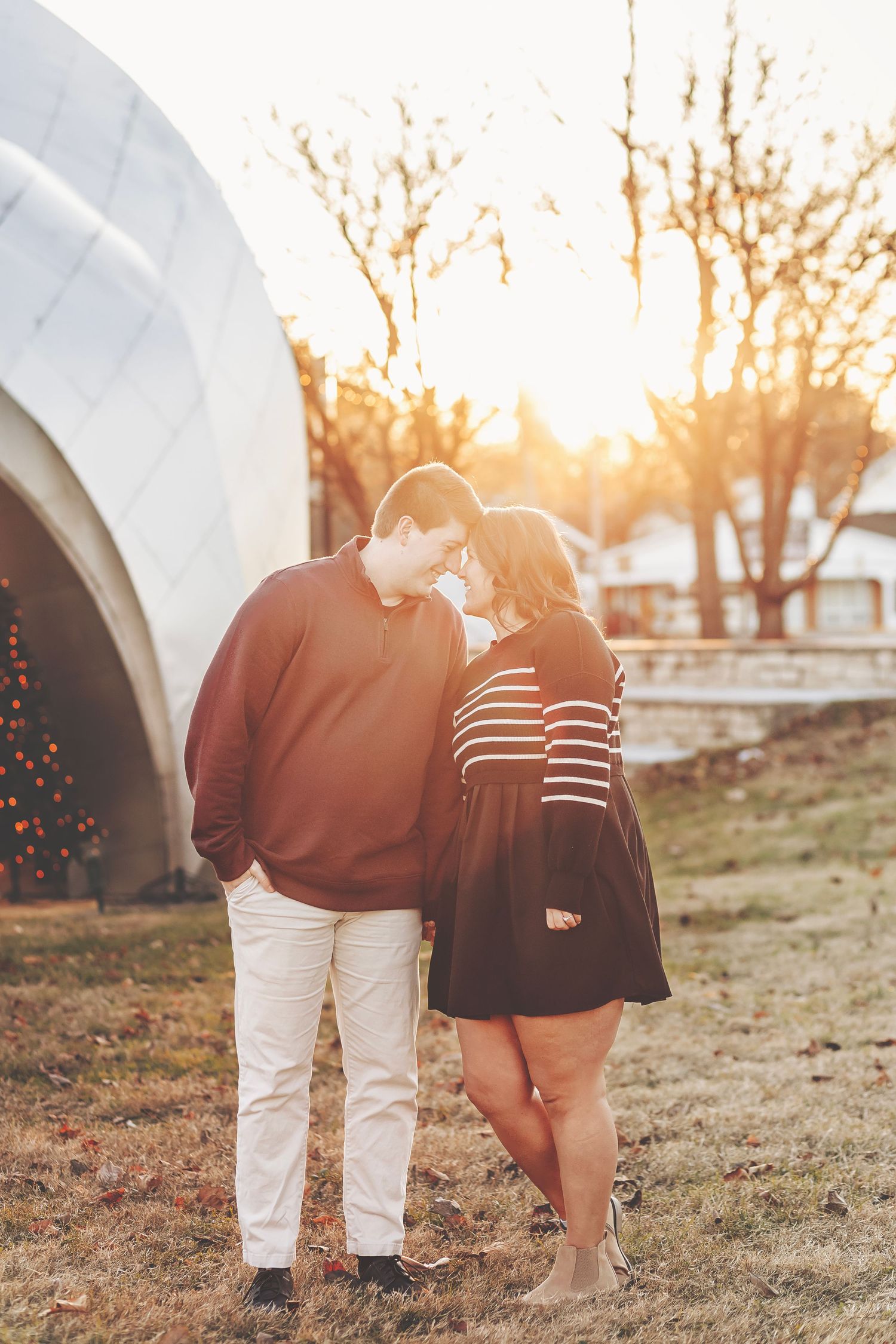 Couple sharing a romantic moment at sunset in a park, man in burgundy sweater and woman in striped black dress.