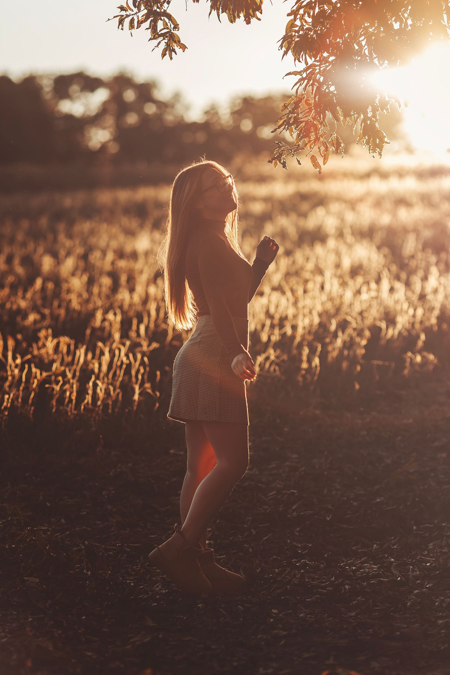 Girl standing in golden field at sunset with warm backlight, long hair, short dress, lens flare, autumn leaves overhead.