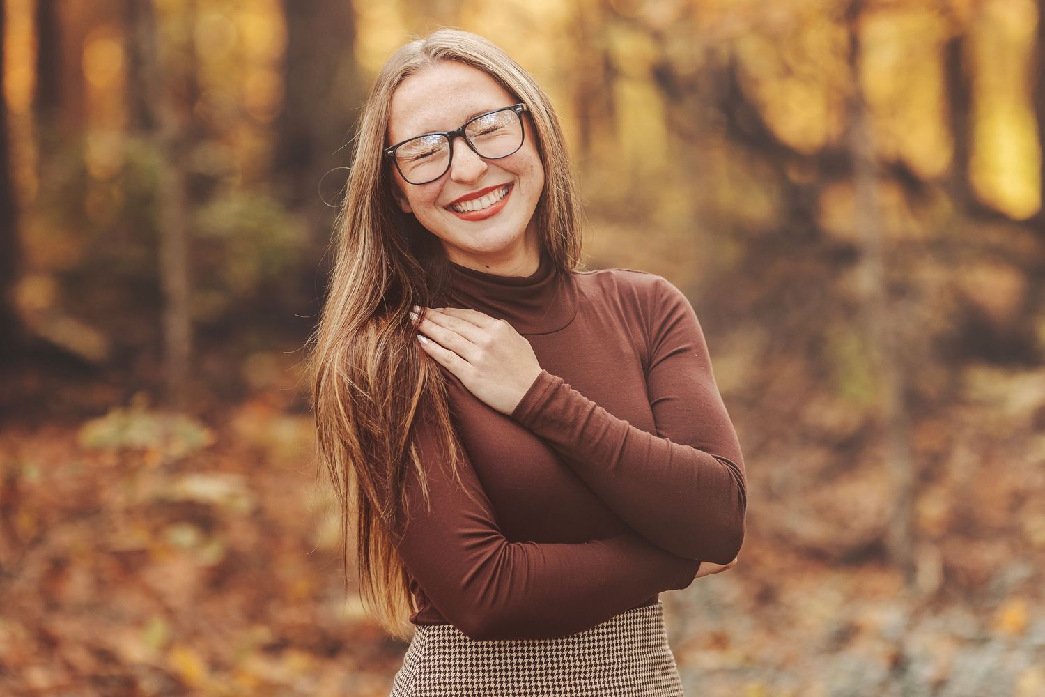 Smiling young woman with glasses in brown turtleneck poses in autumn forest with colorful fall foliage.