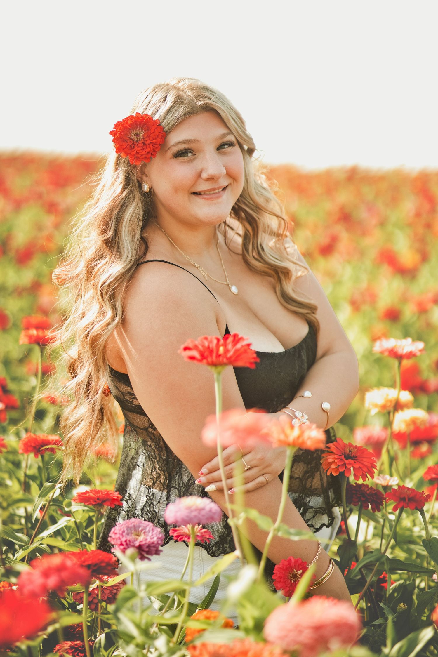 Young woman with red flower in blonde hair sits smiling in a vibrant field of colorful zinnia flowers.