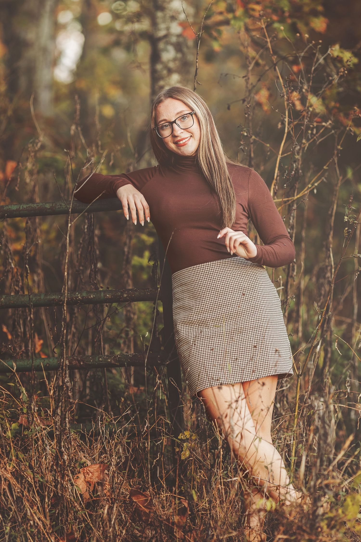 Young woman in brown turtleneck and plaid skirt leaning against old fence in fall foliage.