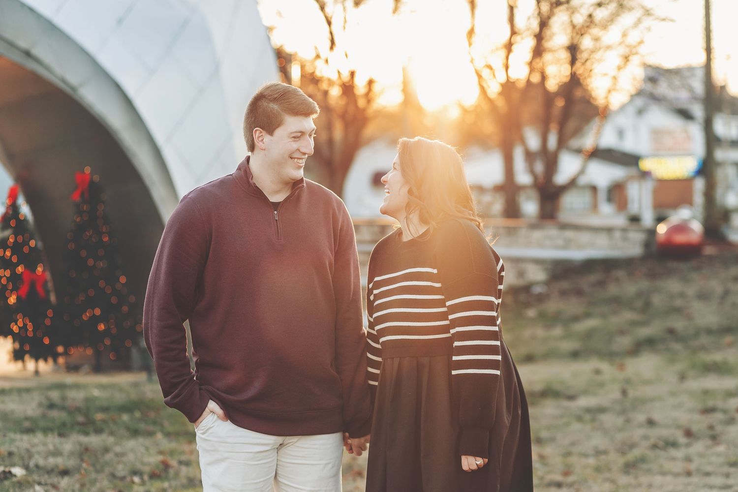Couple holding hands at golden hour sunset outdoors near holiday decorations, man in burgundy sweater, woman in striped dress.