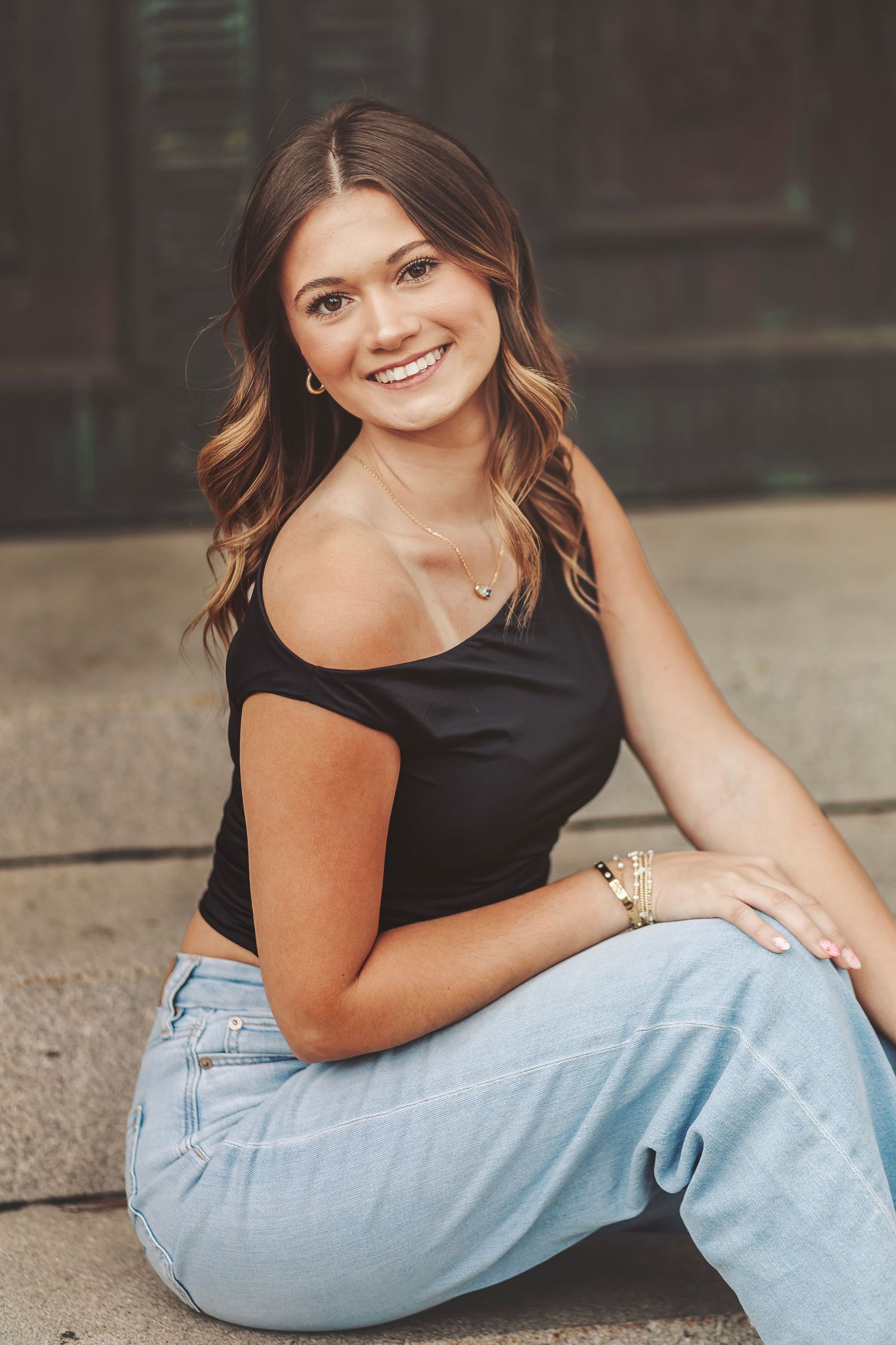 Young woman with wavy brown hair smiling, wearing black crop top and light jeans, sitting outdoors in a relaxed pose.