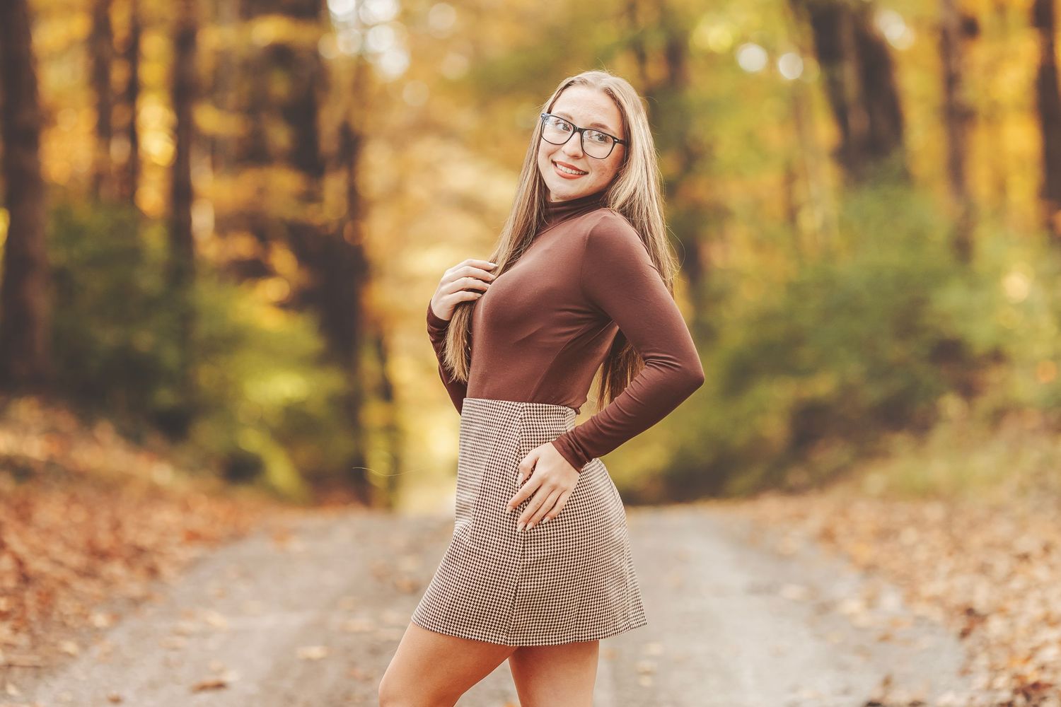 Young woman in brown turtleneck and plaid mini skirt poses on autumn forest path wearing glasses.