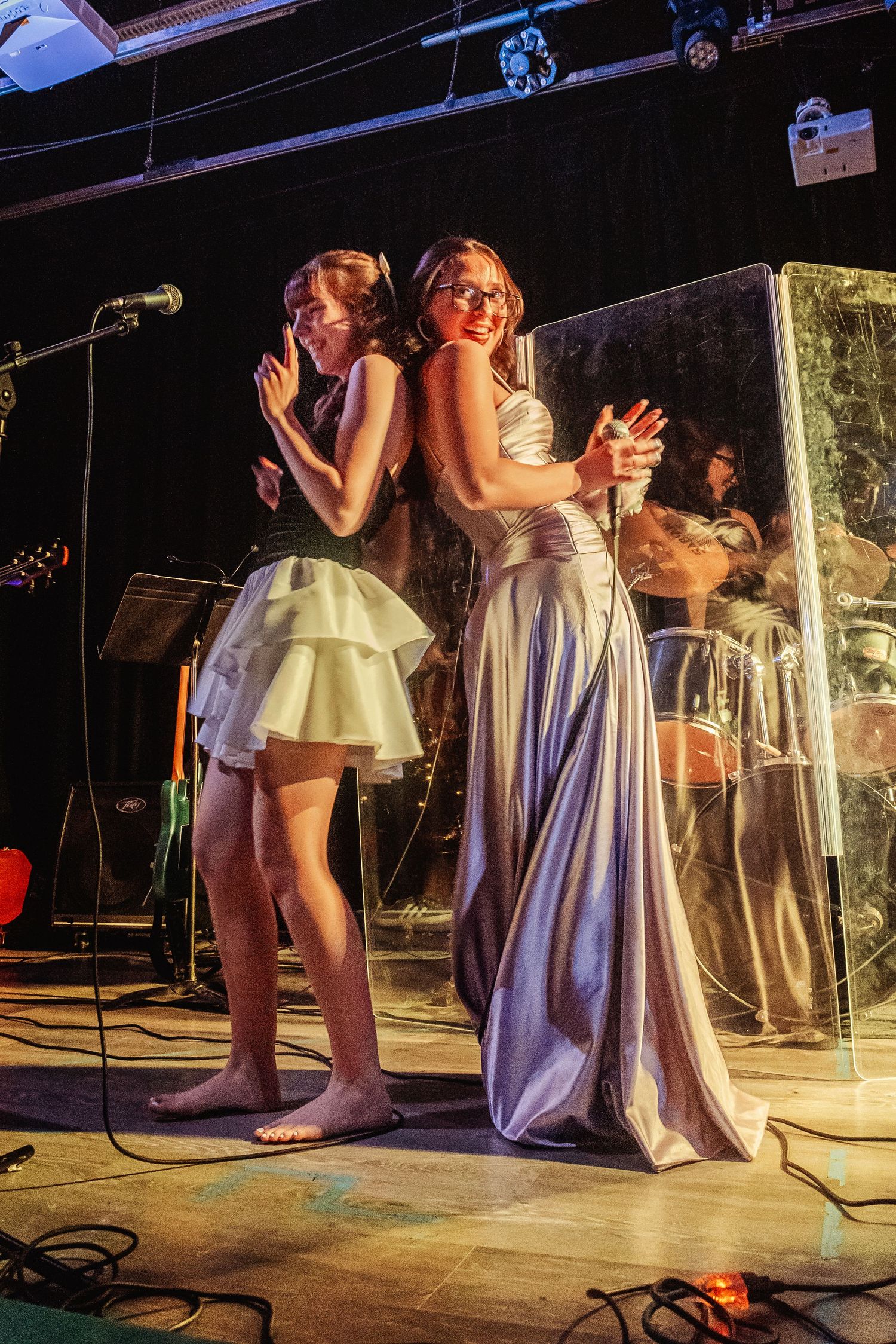 Two young women in elegant dresses performing on stage with microphones at a live music event.