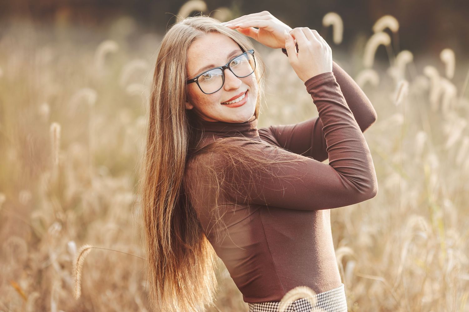 Young woman with long hair and glasses smiling in a sunlit field, wearing a brown turtleneck top.