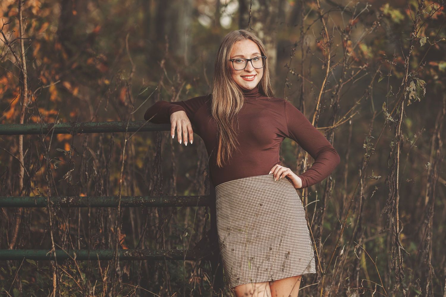 Young woman in glasses, brown turtleneck, and tan skirt poses by a rustic gate surrounded by autumn foliage.