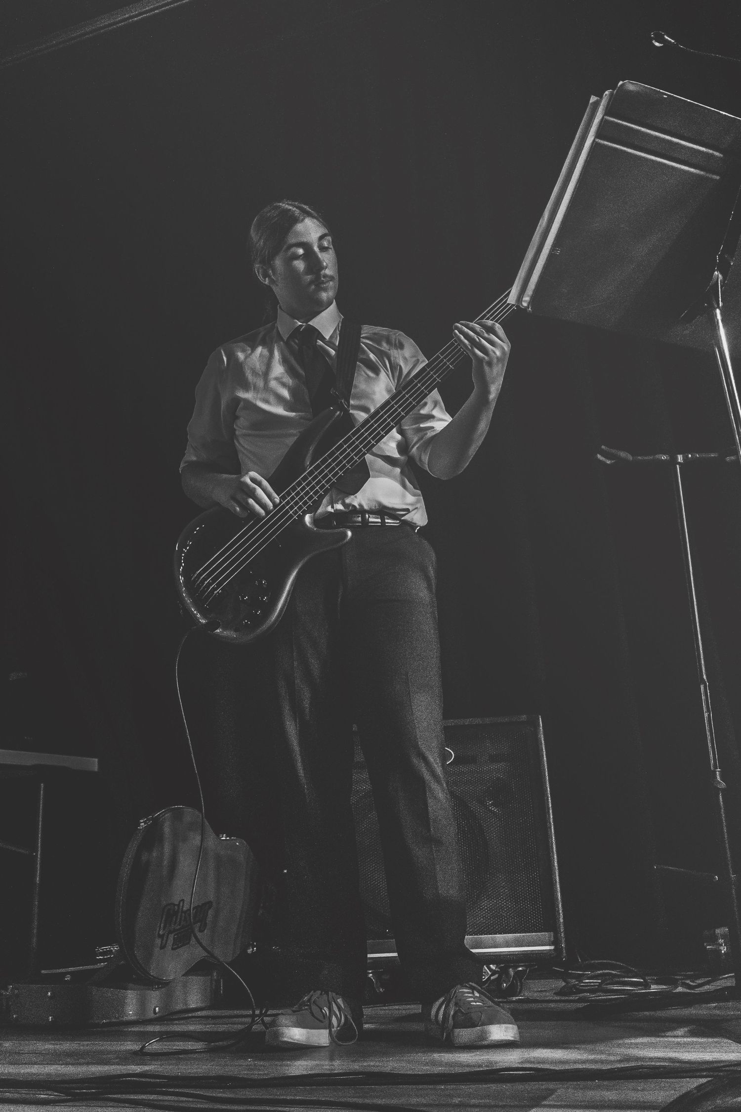 Black and white photo of a male bassist performing on stage in formal attire under dramatic lighting.