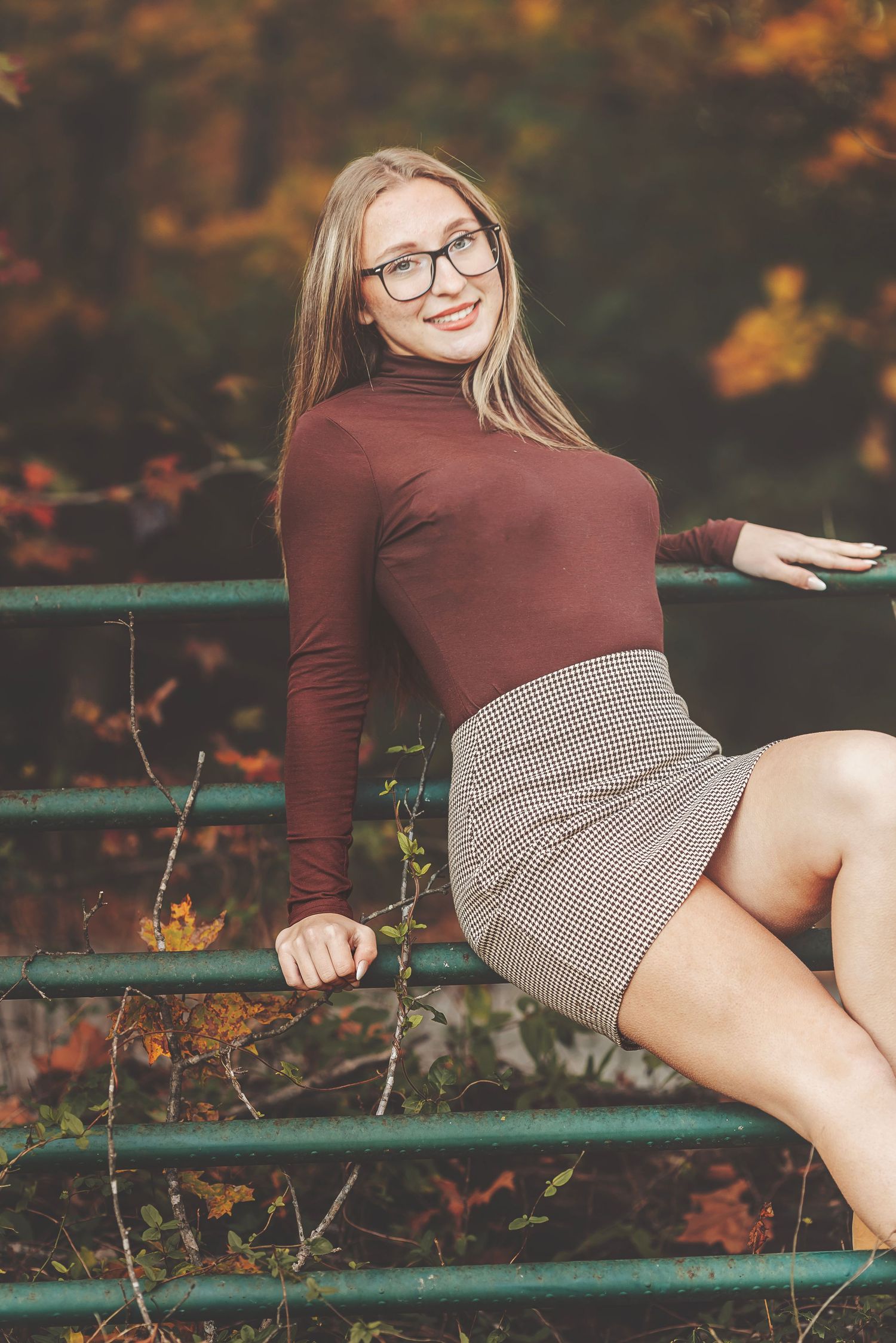 Young blonde woman in glasses, maroon turtleneck, and plaid skirt posing on a fence in an autumn park.