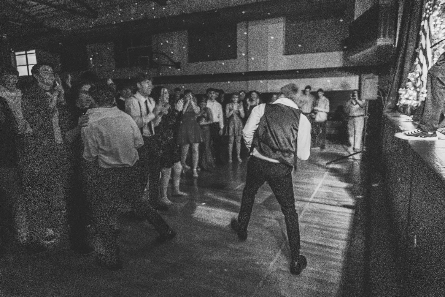 Black and white photo of guests dancing at a lively wedding reception in a rustic venue.