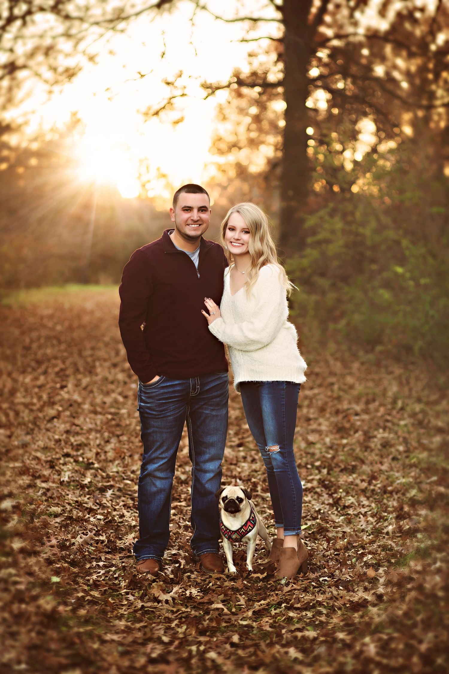 Couple smiling with a pug dog on a leaf-covered forest path during golden hour in autumn.