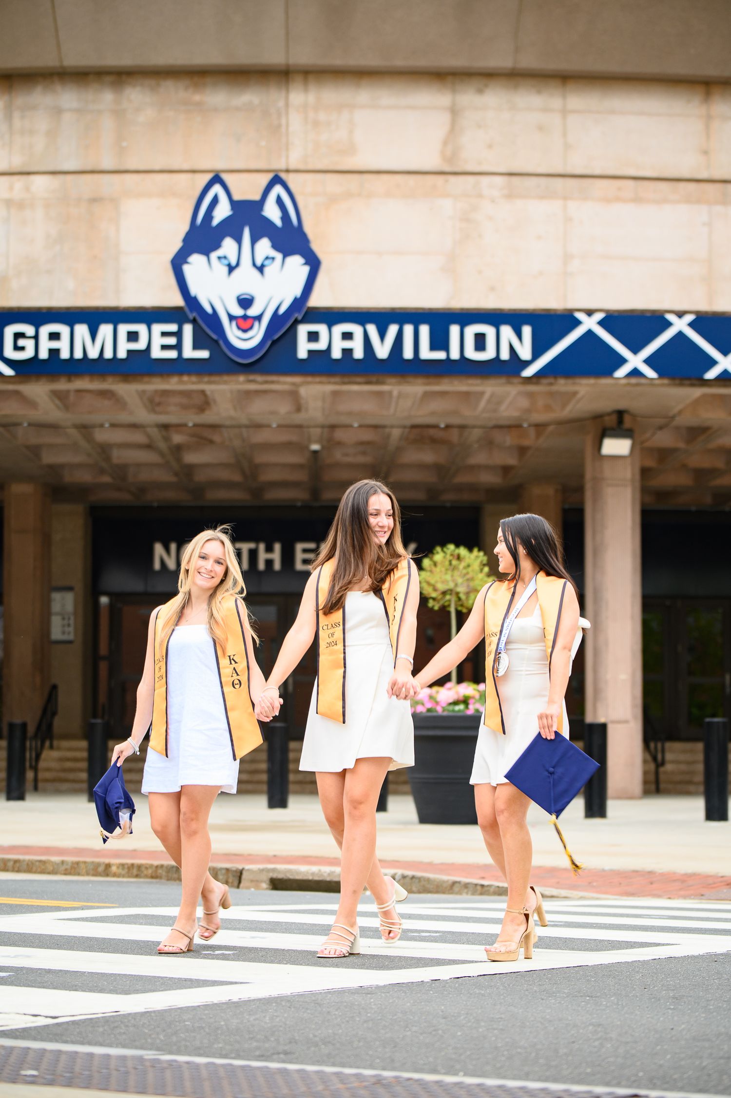 Three people in white dresses and beige sweaters walk together outside the Gampel Pavilion at UConn campus.