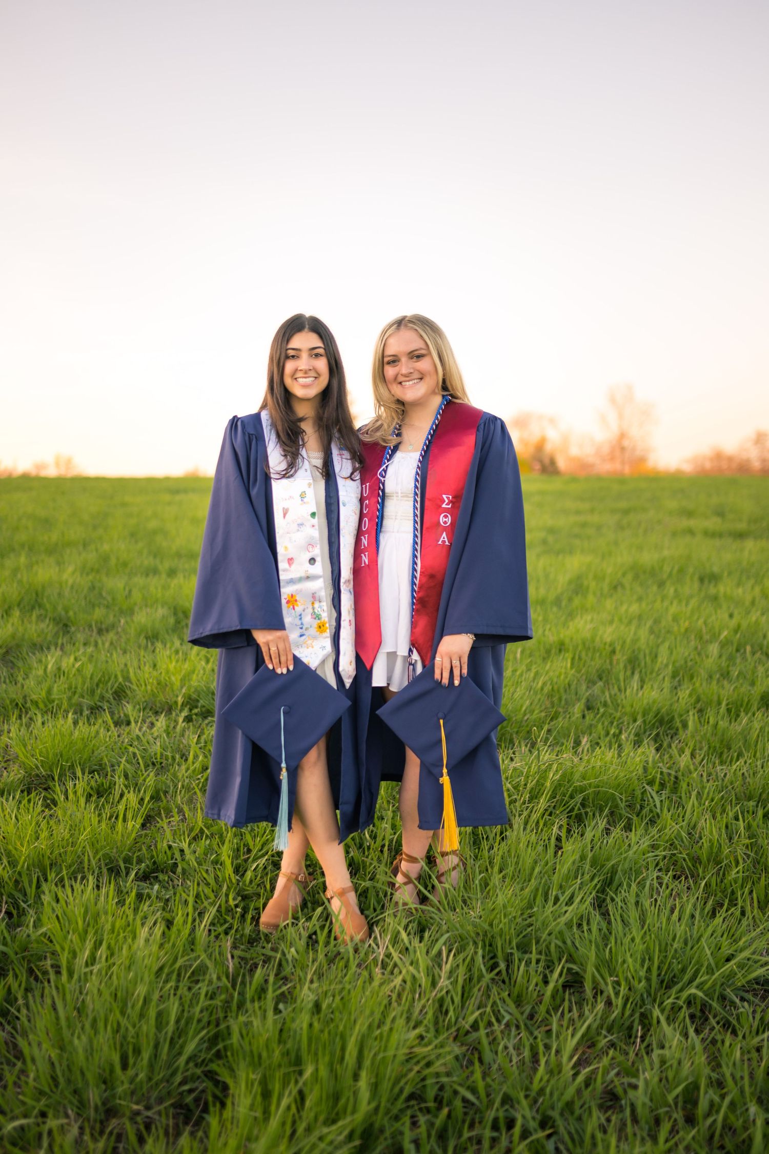 Two graduates wearing navy blue caps and gowns stand together in a green grass field at sunset.