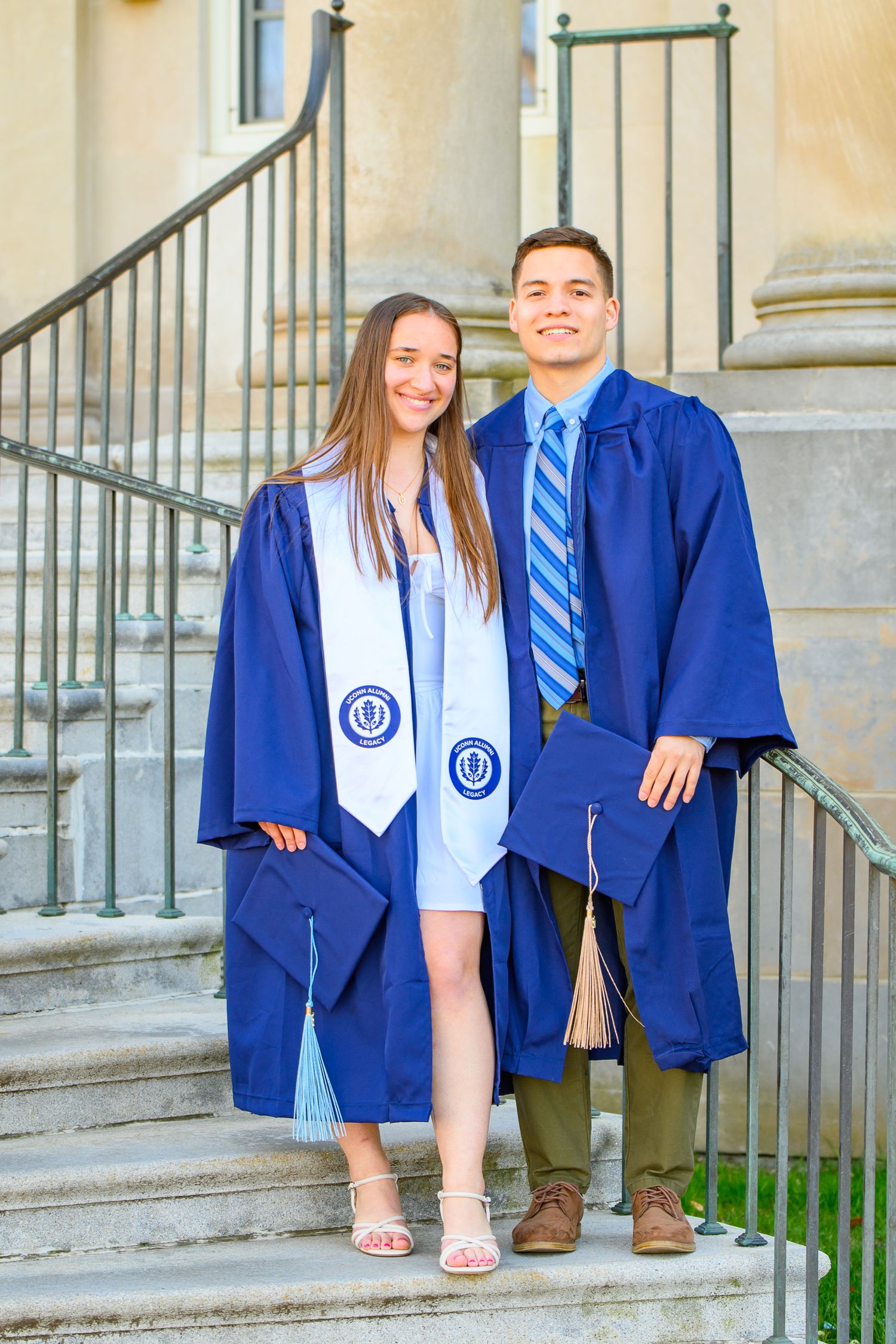 Two graduates in blue caps and gowns stand together on stone steps, wearing matching sashes and graduation attire.
