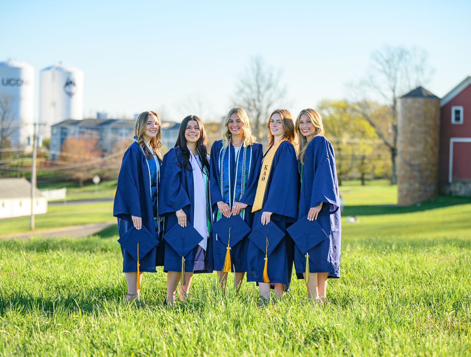 A group in blue graduation gowns stands together on green grass with farm buildings and silos in the background.
