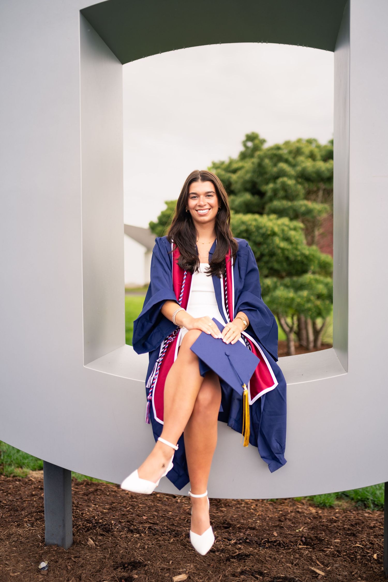 Graduate wearing blue and red academic regalia sits in white architectural alcove during outdoor graduation photoshoot.
