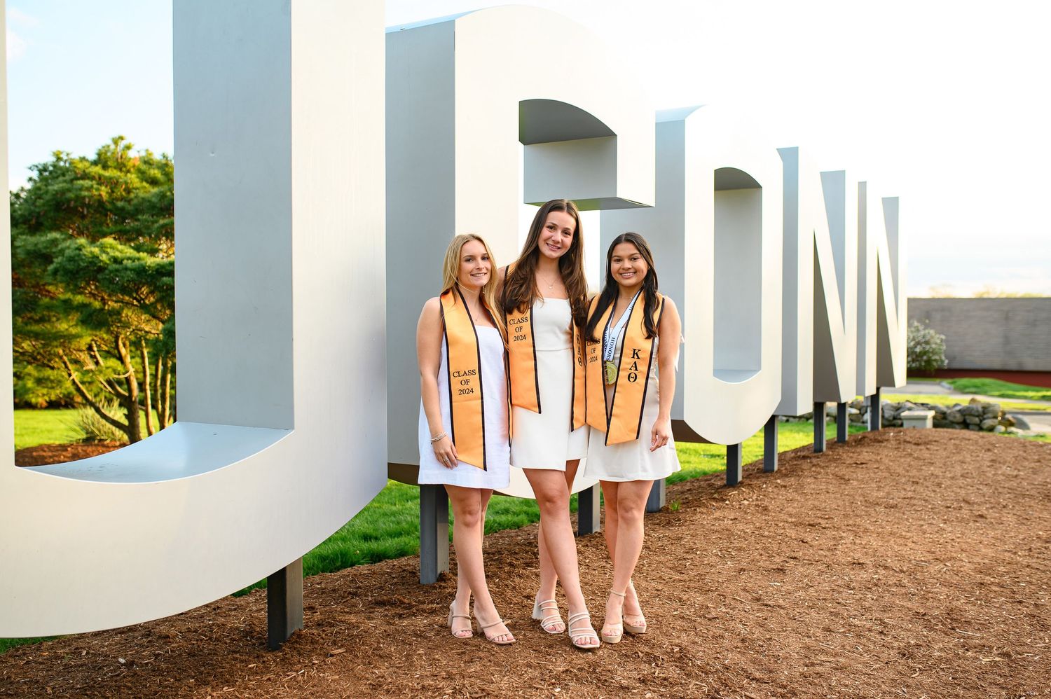 Three graduates in white dresses and gold stoles pose together in front of large decorative letters on campus grounds.
