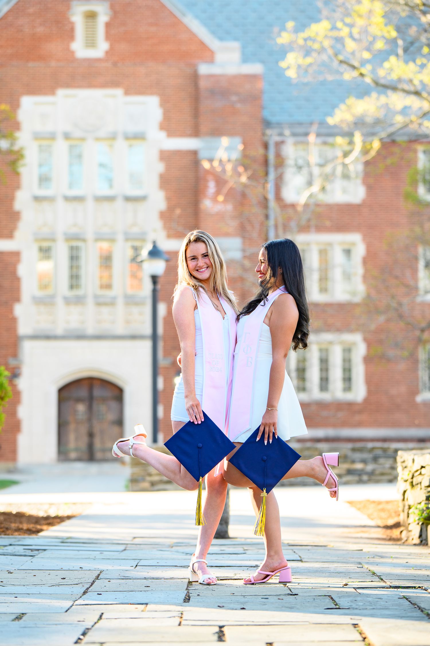 Two graduates in dresses holding diplomas pose playfully on a stone path in front of a historic brick university building.